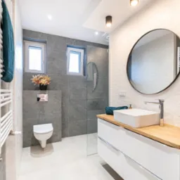 Nowoczesna łazienka w Katowicach z szarymi płytkami, owalnym lustrem, białą umywalką nablatową na drewnianym blacie i grzejnikiem drabinkowym z turkusowym ręcznikiem.