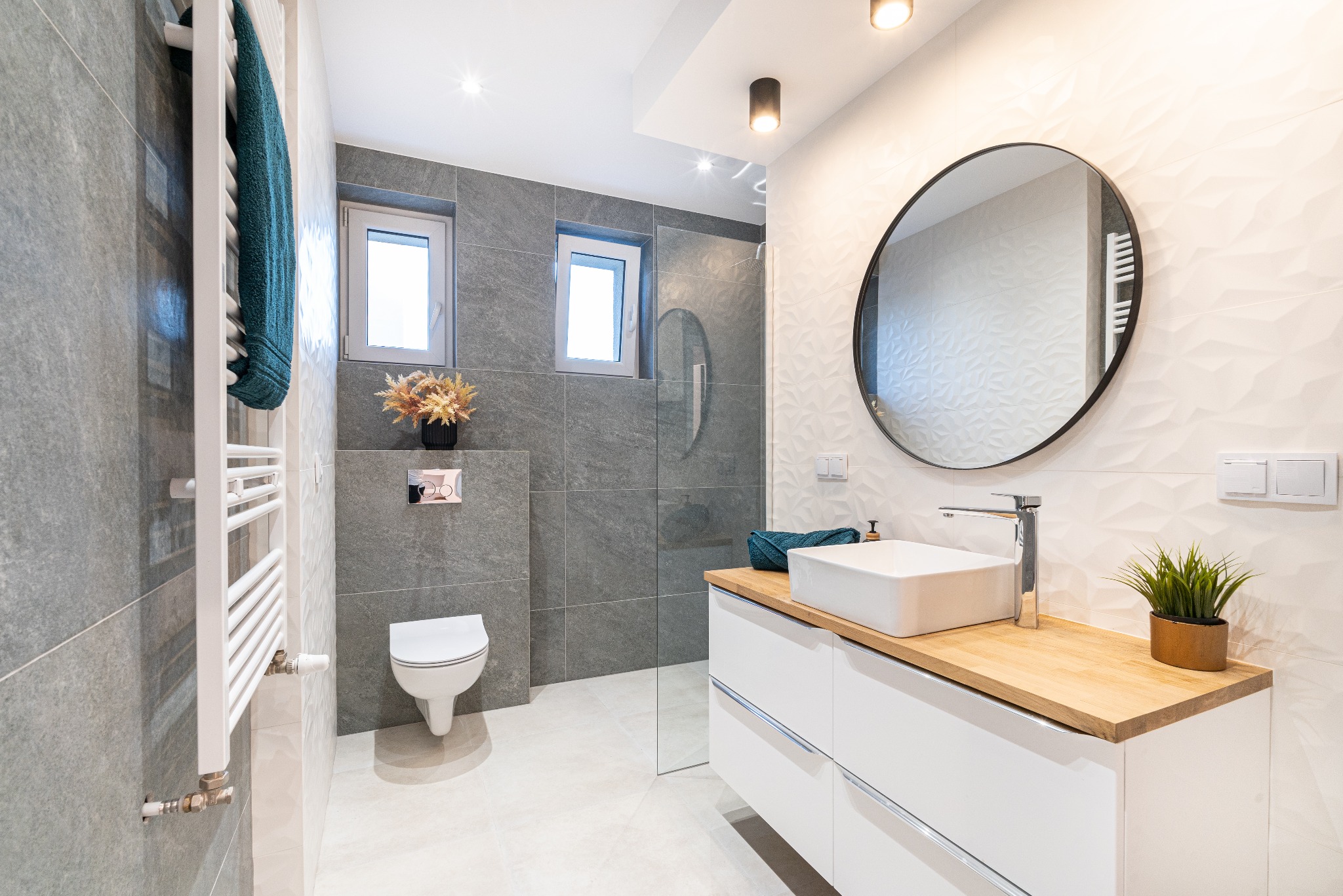Nowoczesna łazienka w Katowicach z szarymi płytkami, owalnym lustrem, białą umywalką nablatową na drewnianym blacie i grzejnikiem drabinkowym z turkusowym ręcznikiem.
