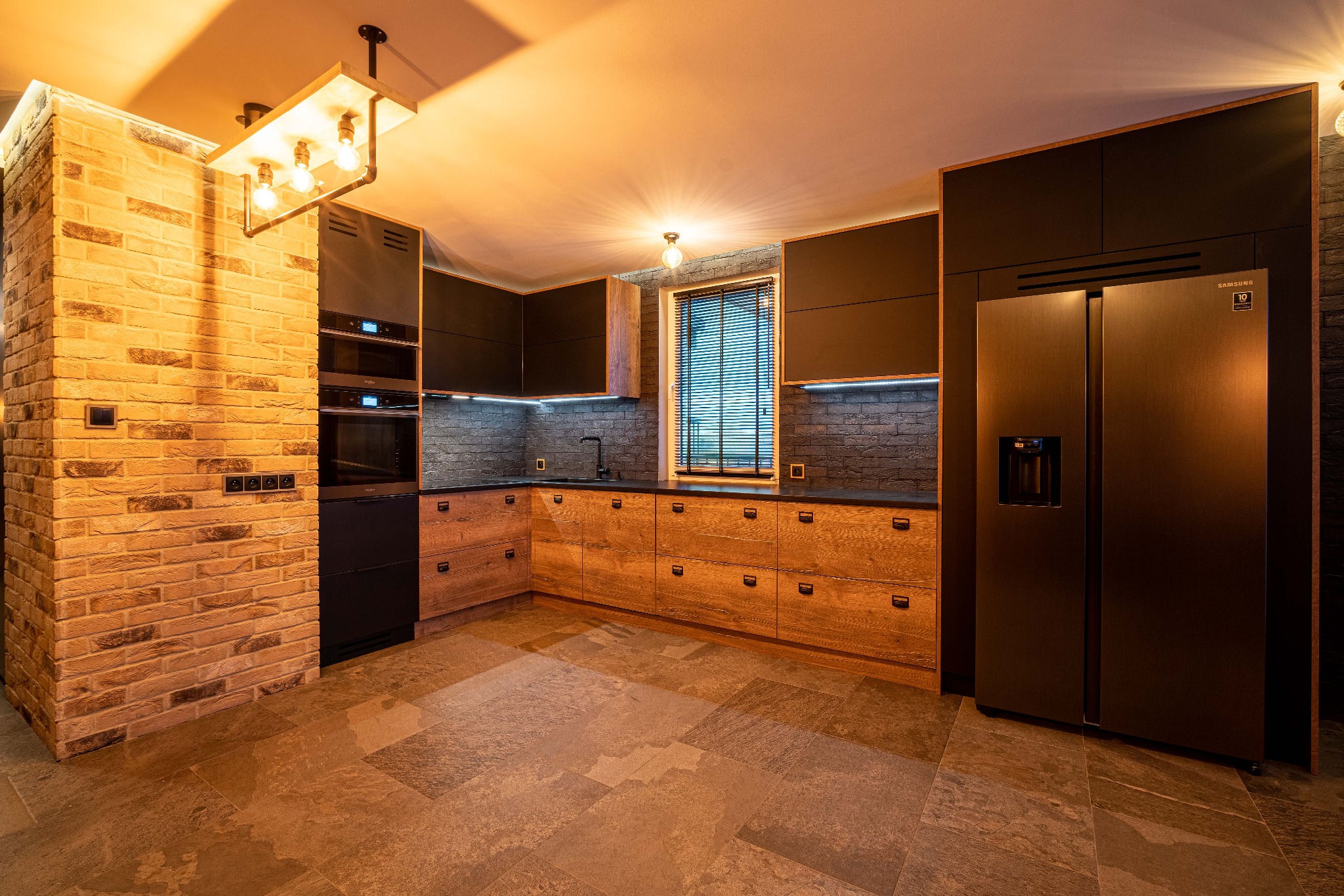 Nowoczesna kuchnia z ceglaną ścianą, drewnianymi szafkami i czarnymi elementami, oświetlona ciepłym światłem, z widoczną lodówką Samsung.