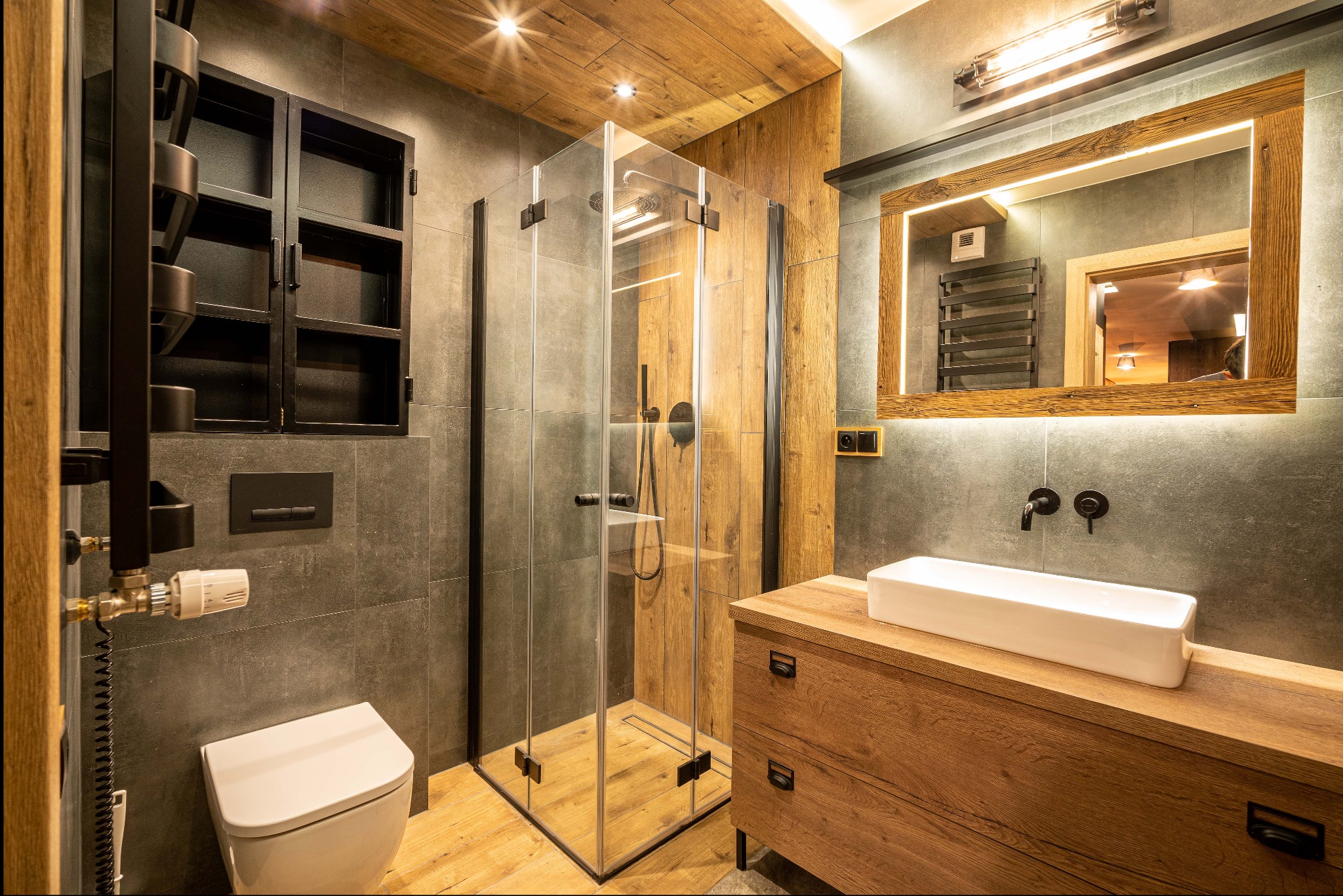 Nowoczesna łazienka z kabiną prysznicową typu walk-in, umywalką nablatową na drewnianej szafce, podświetlanym lustrem w drewnianej ramie i czarnymi elementami armatury.