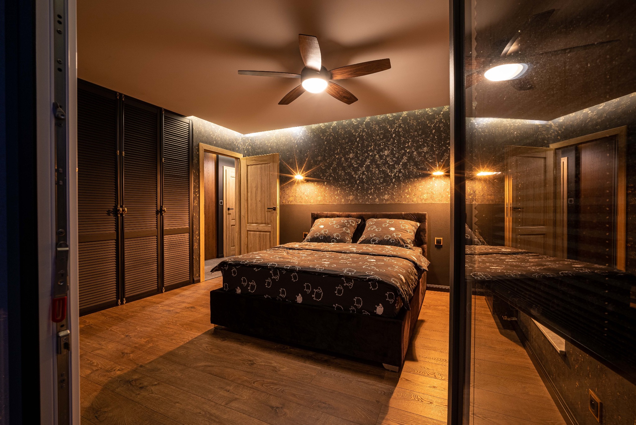 Sypialnia z łóżkiem małżeńskim, drewnianym wentylatorem sufitowym, szafą z żaluzjowymi drzwiami i dekoracyjną tapetą w odcieniach złota, widoczna przez otwarte drzwi.