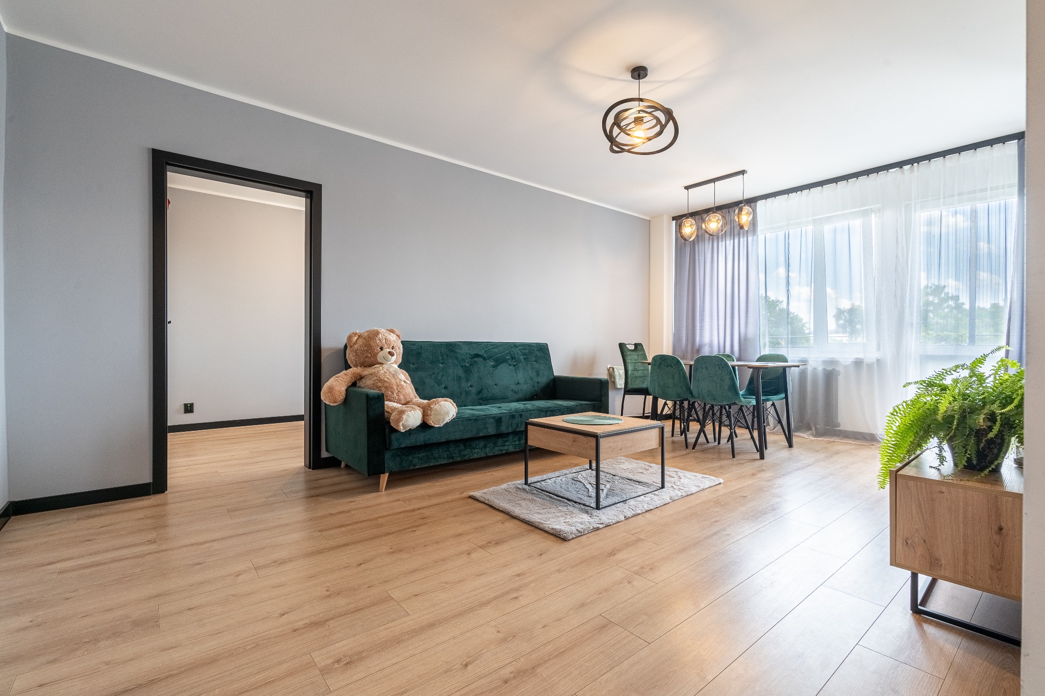Jasne wnętrze pokoju z zieloną sofą, na której siedzi pluszowy miś, stół z krzesłami, drewniana podłoga i szafka z rośliną doniczkową.