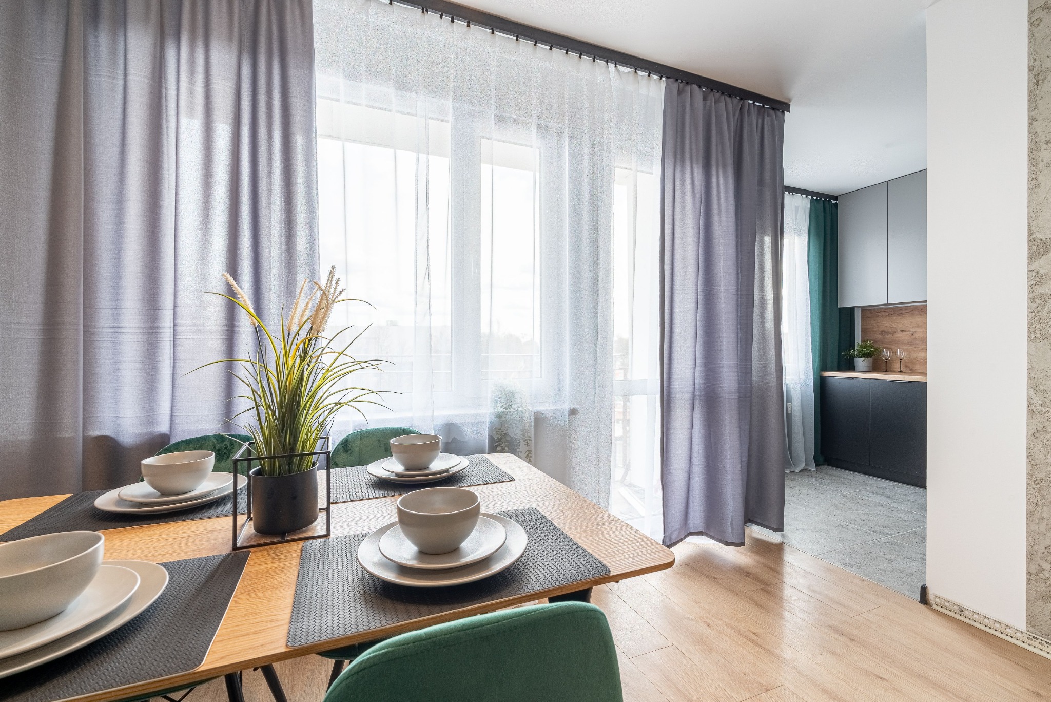 Jasna jadalnia z drewnianym stołem nakrytym dla dwóch osób, zielone krzesła, dekoracyjna trawa w czarnej doniczce, okno z szarymi i białymi zasłonami, widok na kuchnię w tle.