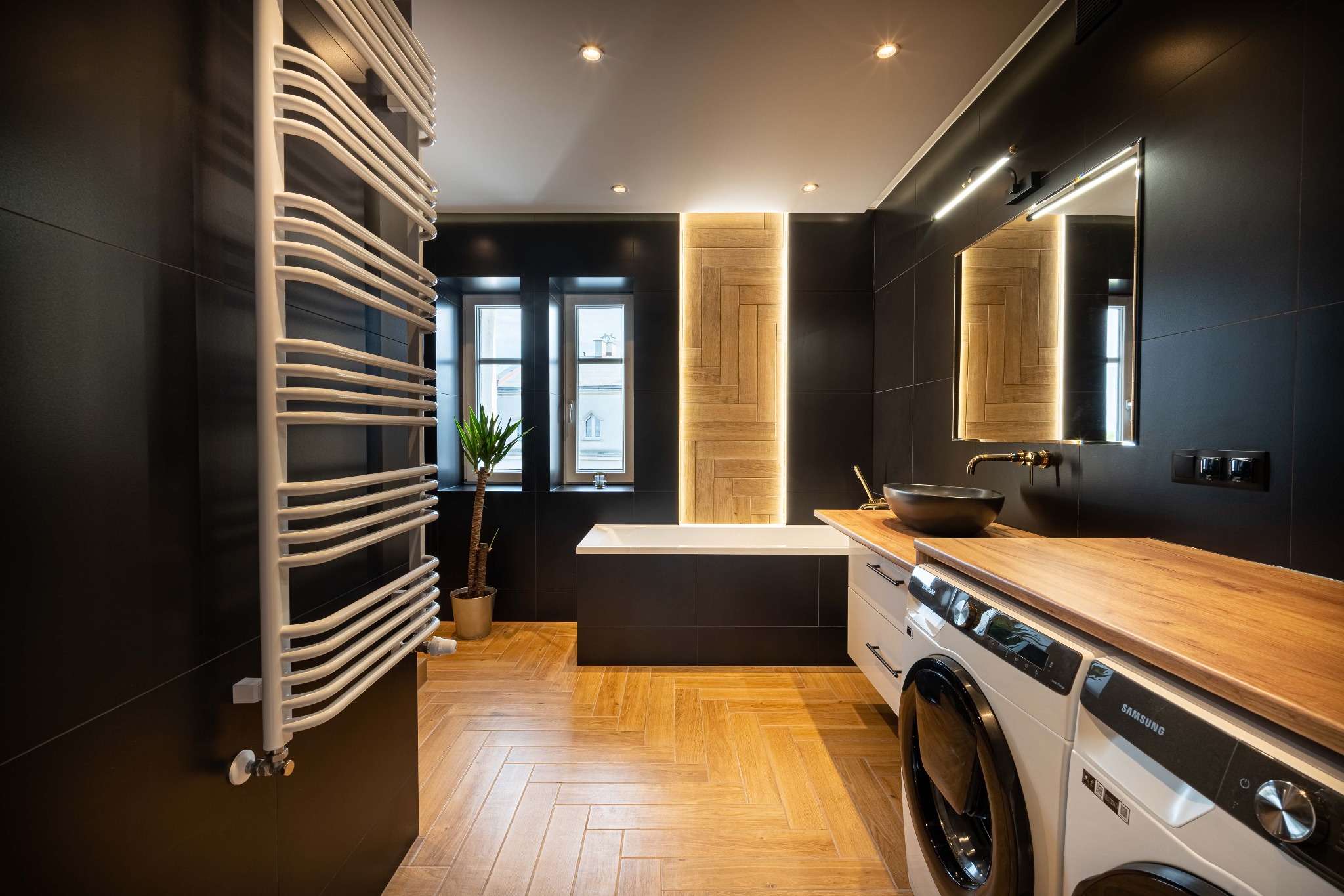 Nowoczesna łazienka z czarnymi płytkami, drewnianą podłogą w jodełkę, grzejnikiem drabinkowym, wanną, umywalką nablatową i pralką z drewnianym blatem.
