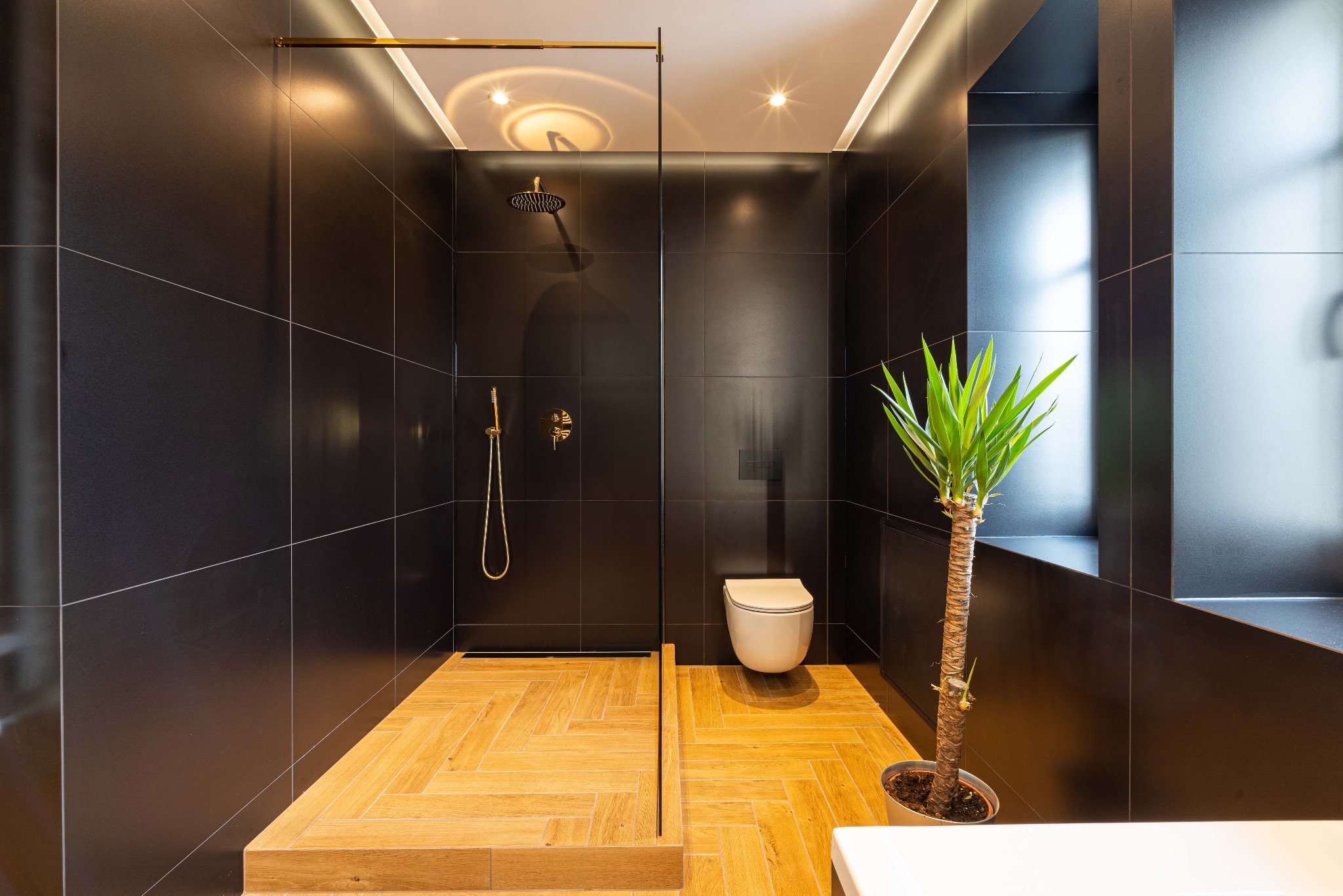Nowoczesna łazienka z czarnymi płytkami, drewnopodobną podłogą w jodełkę, szklaną kabiną prysznicową z złotą armaturą i rośliną doniczkową.
