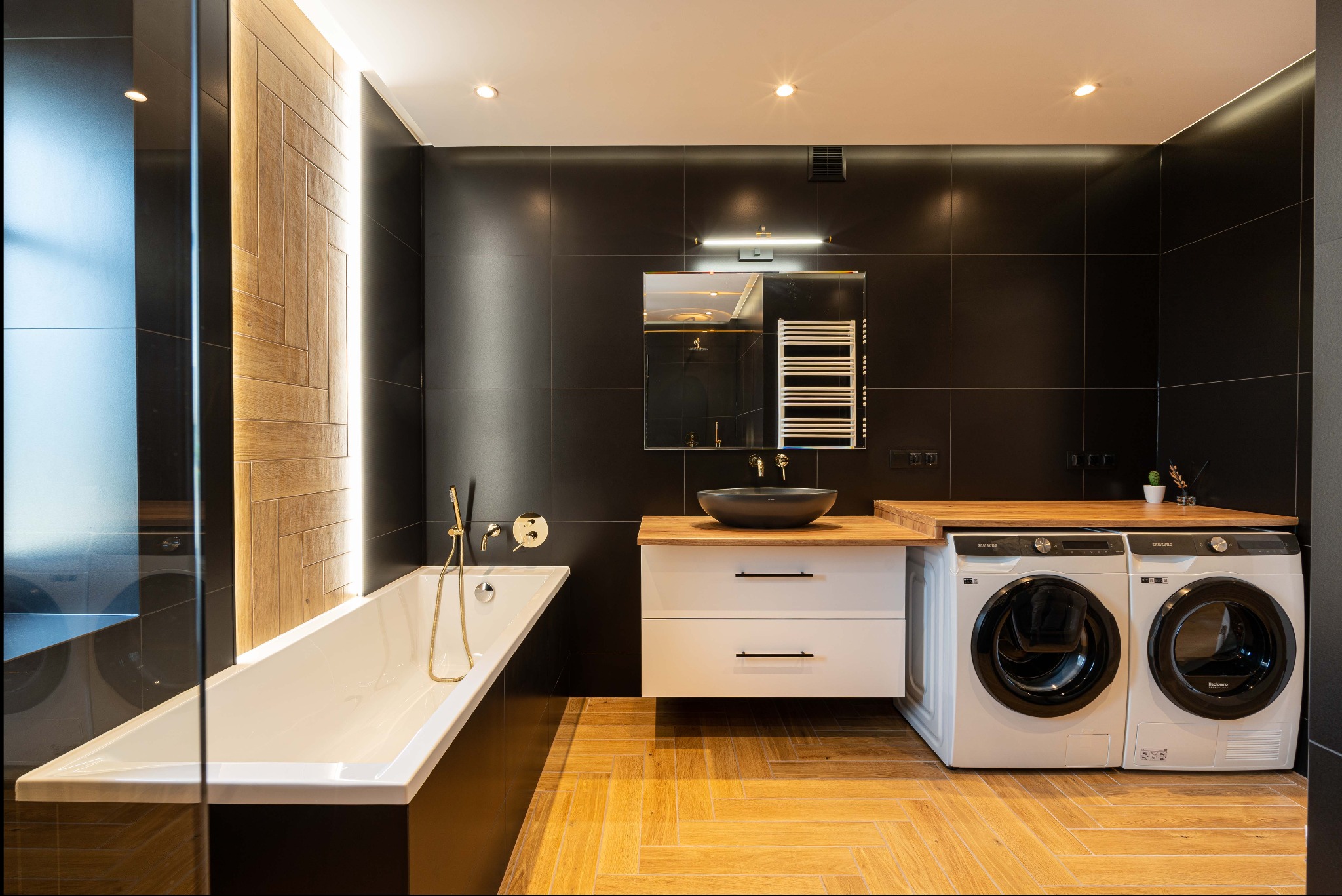 Nowoczesna łazienka z czarnymi płytkami, drewnianą podłogą ułożoną w jodełkę, białą wanną, umywalką nablatową i pralko-suszarką Samsung.