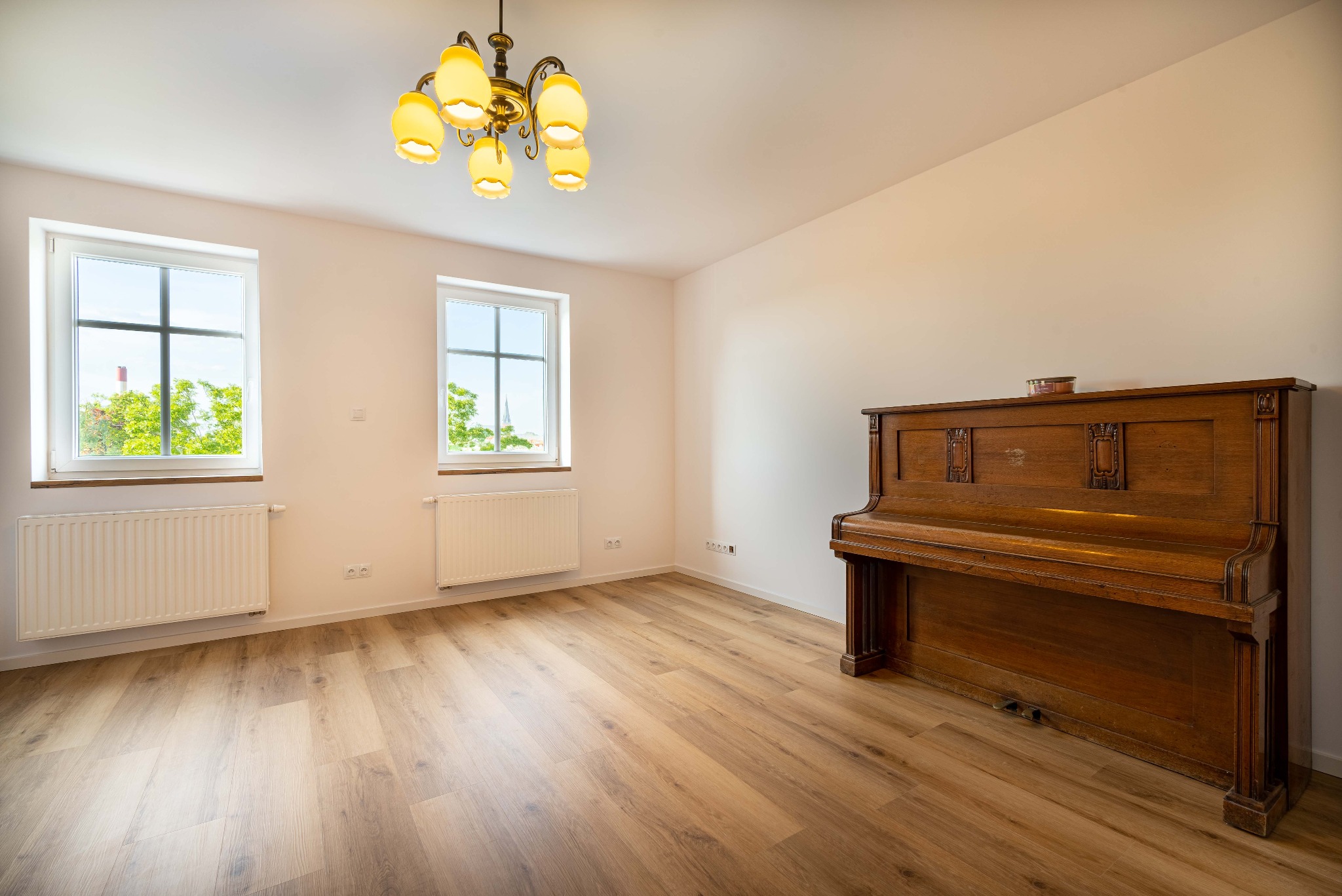 Jasne, puste pomieszczenie z drewnianą podłogą, dwoma oknami z widokiem na zieleń i zabytkowym pianinem przy ścianie, oświetlone ozdobnym, żółtym żyrandolem.