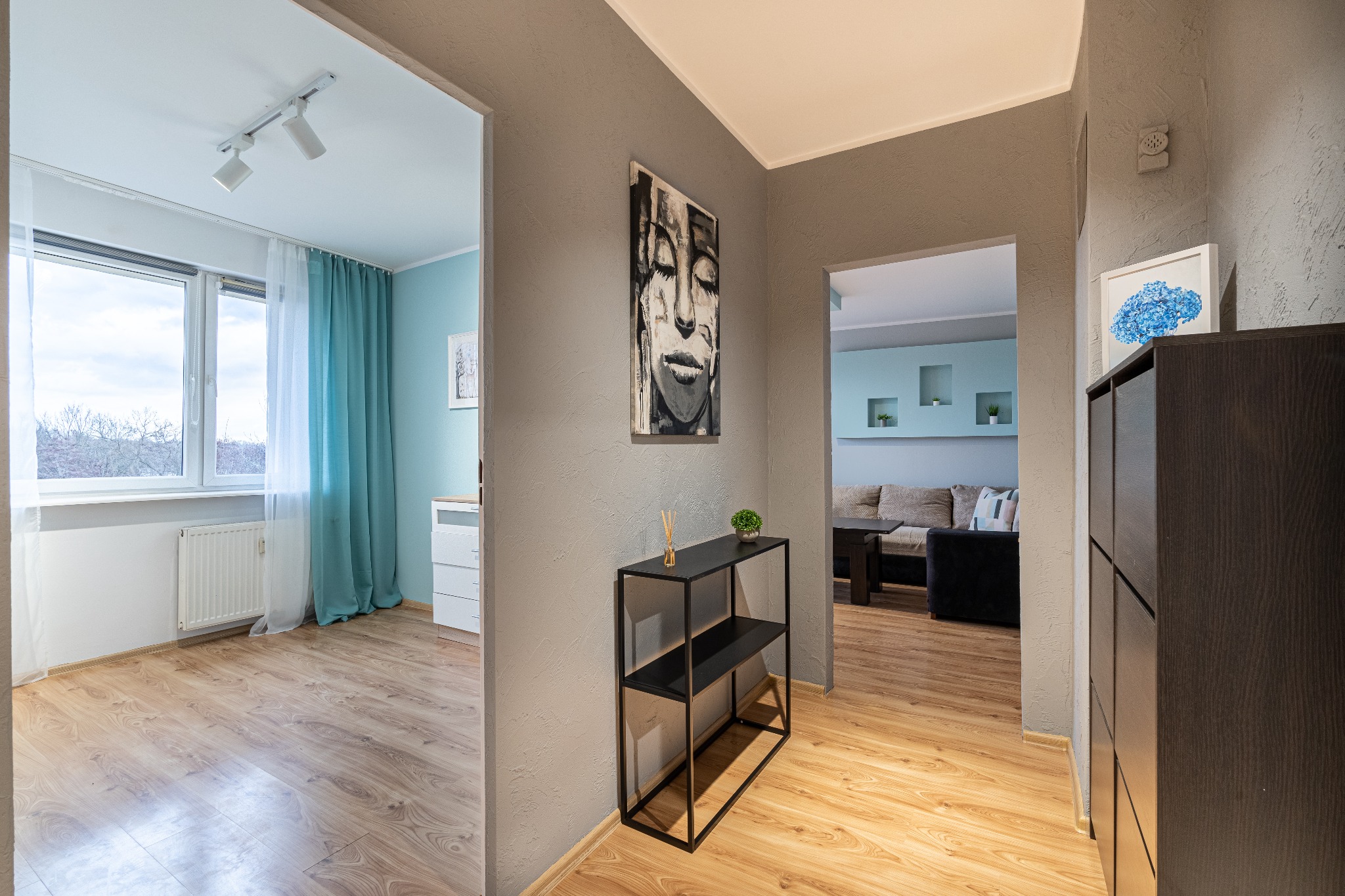 Wnętrze mieszkania: korytarz z widokiem na salon i pokój, szara ściana z abstrakcyjnym obrazem kobiety, czarny regał z dekoracjami, parkiet, szafa wenge, niebieska hortensja w ramce.