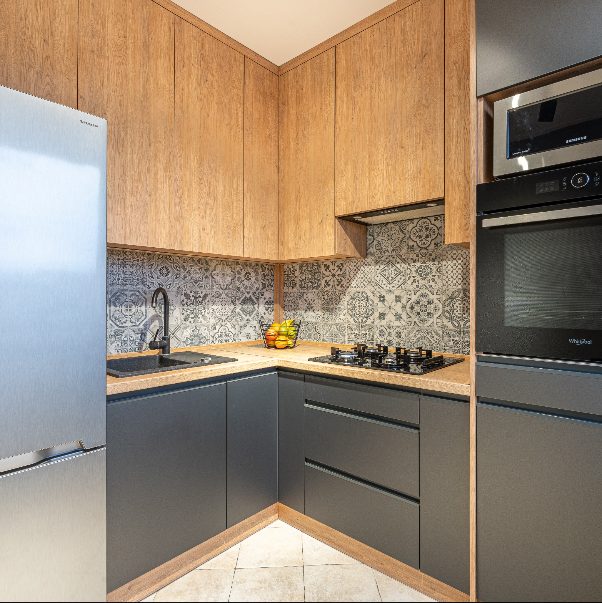 Nowoczesna kuchnia w kształcie litery L z drewnianymi szafkami górnymi, szarymi dolnymi, czarnym zlewem i baterią, oraz wzorzystymi płytkami na ścianie.