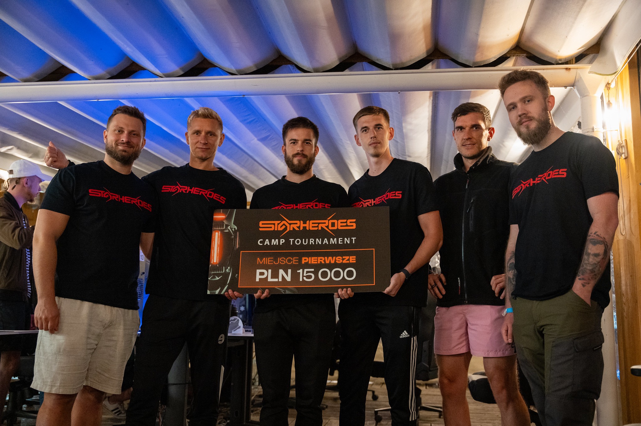 Drużyna E-sportowa StarHeroes prezentuje tablicę z nagrodą za pierwsze miejsce w turnieju, kwota 15 000 PLN, zawodnicy ubrani w czarne koszulki z logo StarHeroes.