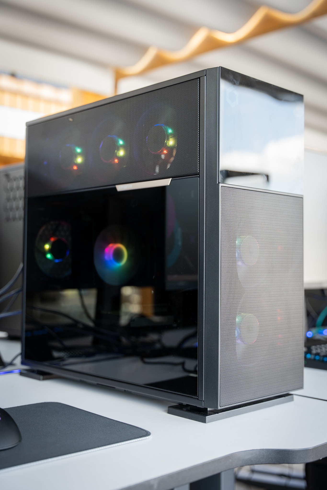 Czarna obudowa komputera PC z przezroczystym panelem bocznym, podświetlanymi wentylatorami RGB i siatkową górą, stojąca na białym biurku obok myszki.