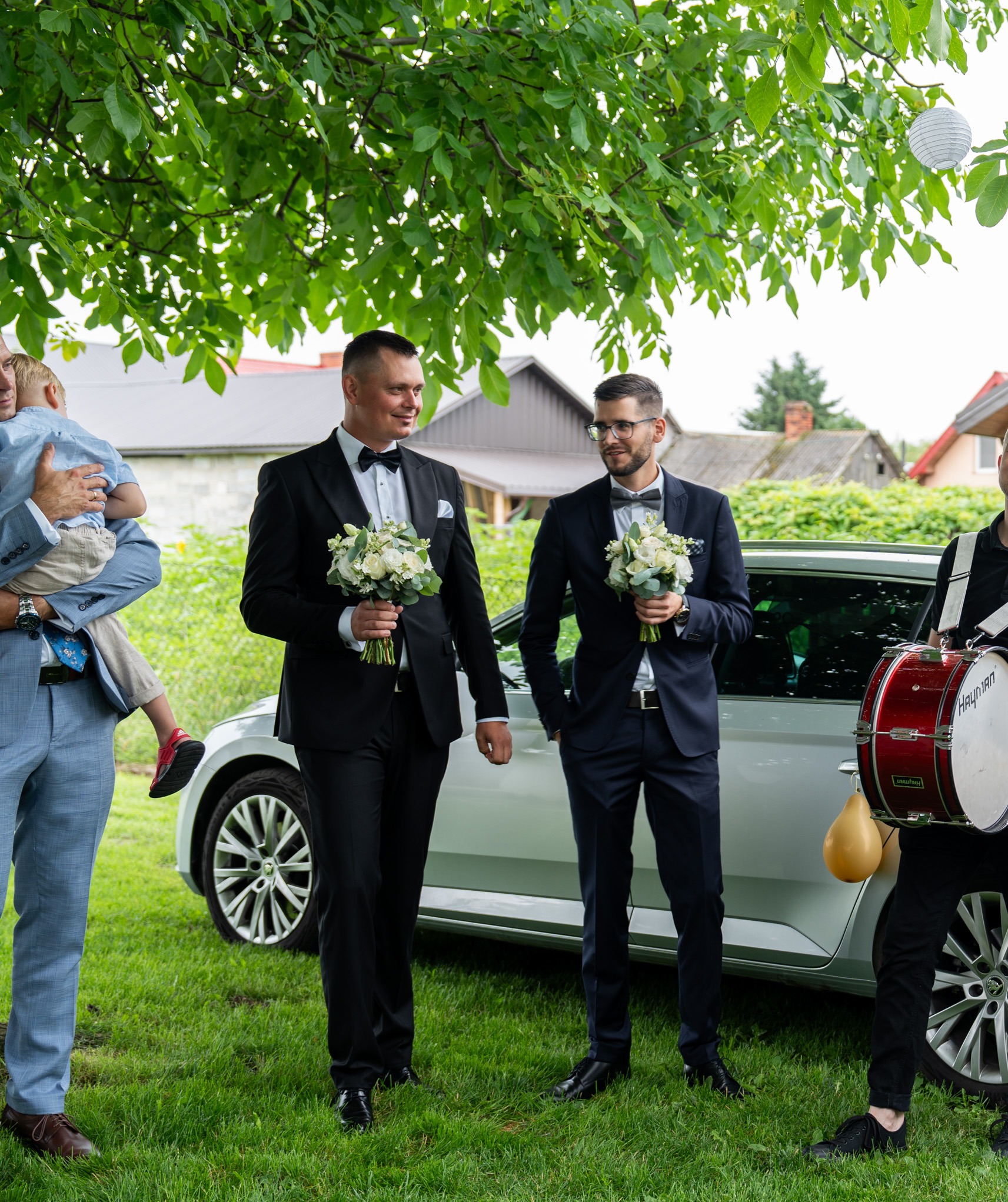Panowie w garniturach z bukietami przed ślubem, jeden z nich trzyma dziecko, w tle samochód i perkusista z bębnem