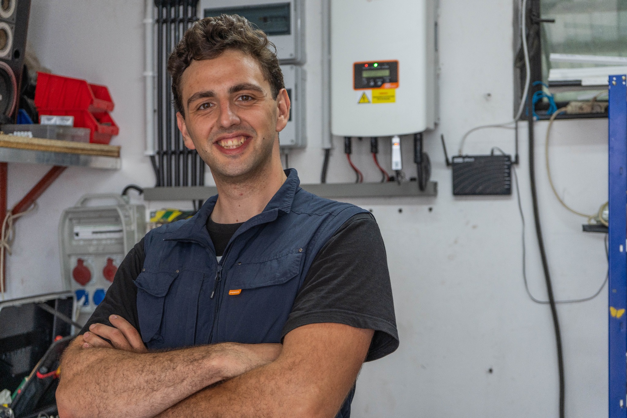 Uśmiechnięty mężczyzna w roboczym ubraniu, z założonymi rękami, na tle ściany z zamontowanymi urządzeniami elektrycznymi i skrzynką narzędziową.