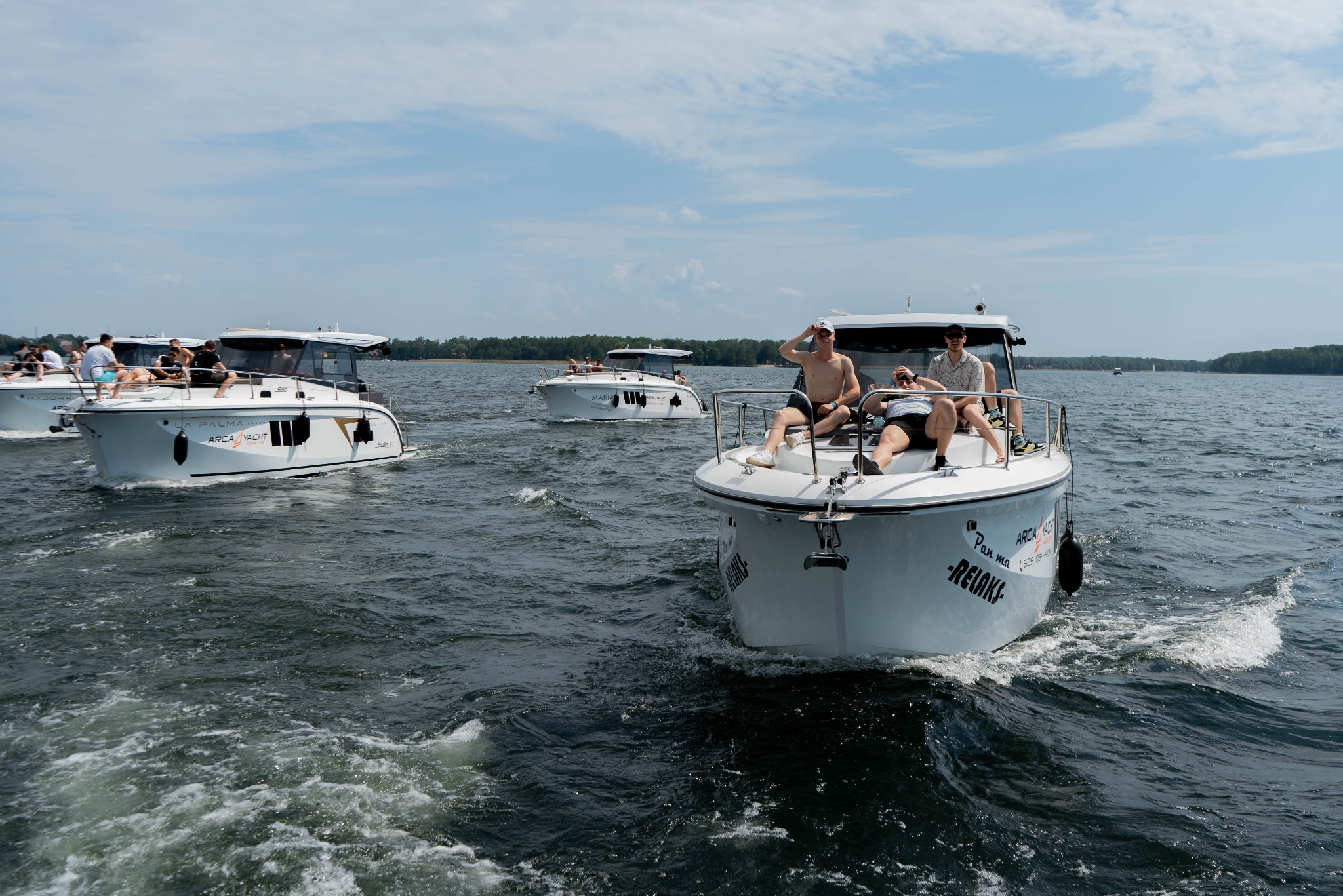 Grupa osób relaksuje się na pokładzie motorowych jachtów podczas słonecznego dnia na jeziorze, z widocznymi falami i bryzą.