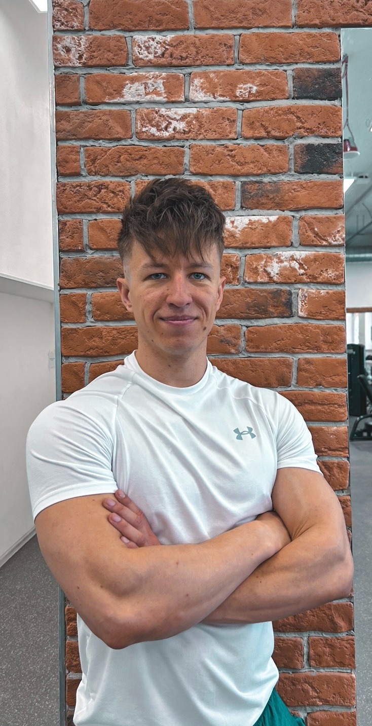 Uśmiechnięty mężczyzna w białej koszulce sportowej z założonymi rękami, oparty o ścianę z czerwonej cegły w siłowni.