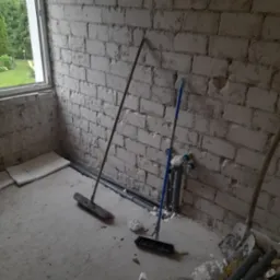 Pok&oacute;j w trakcie remontu: surowe białe cegły, rury przy podłodze, częściowo położone płytki, dwie szczotki i łopata w rogu.