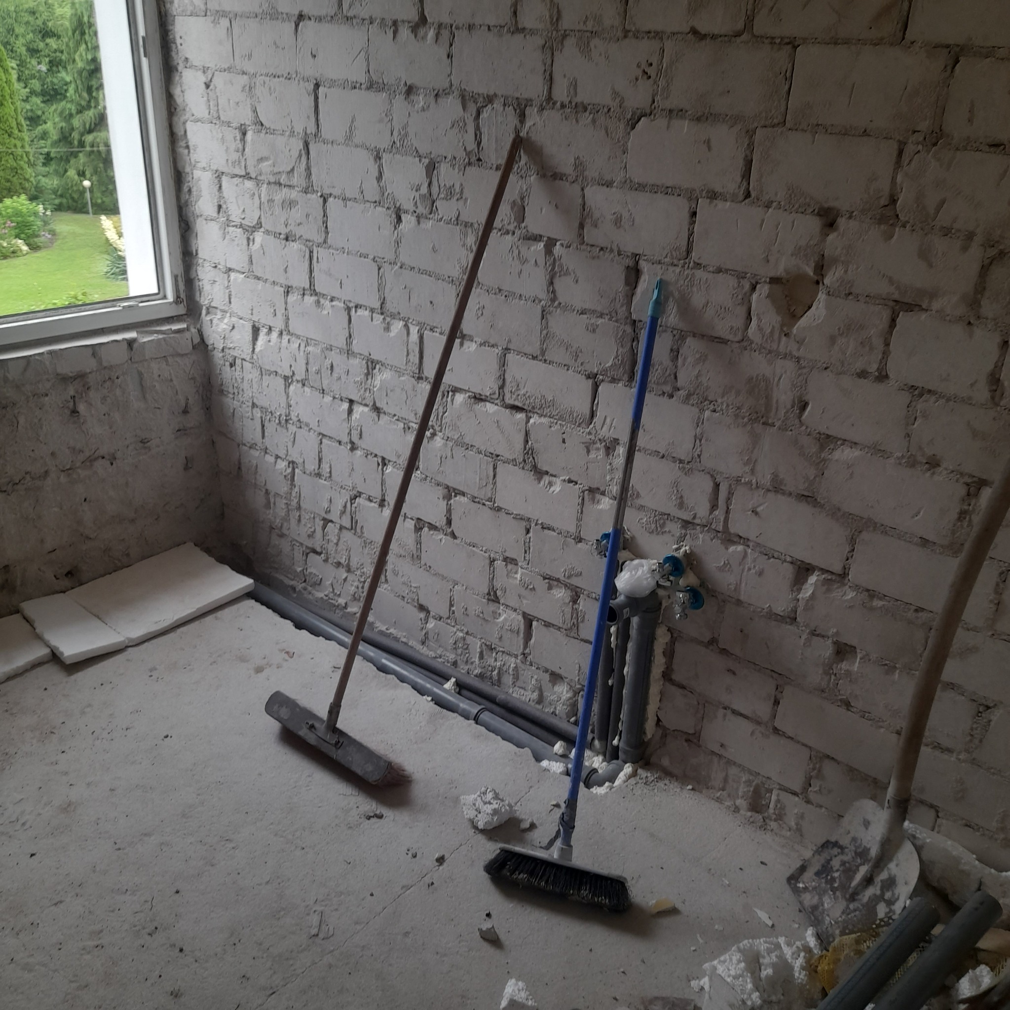 Pokój w trakcie remontu: surowe białe cegły, rury przy podłodze, częściowo położone płytki, dwie szczotki i łopata w rogu.