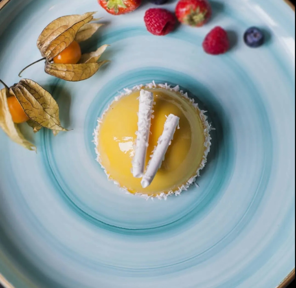 Żółty deser z dwoma białymi dekoracjami na wierzchu, otoczony wiórkami kokosowymi, podany na turkusowym talerzu z owocami w tle