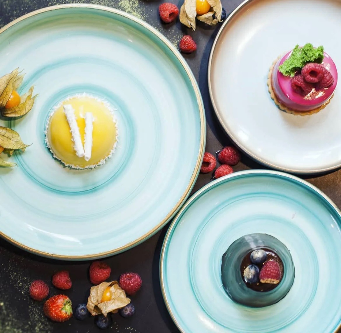 Trzy talerze z deserami: żółty z białymi paskami, różowy z malinami i zielonymi listkami, oraz ciemny z borówkami i maliną, otoczone świeżymi owocami i posypane zielonym proszkiem.