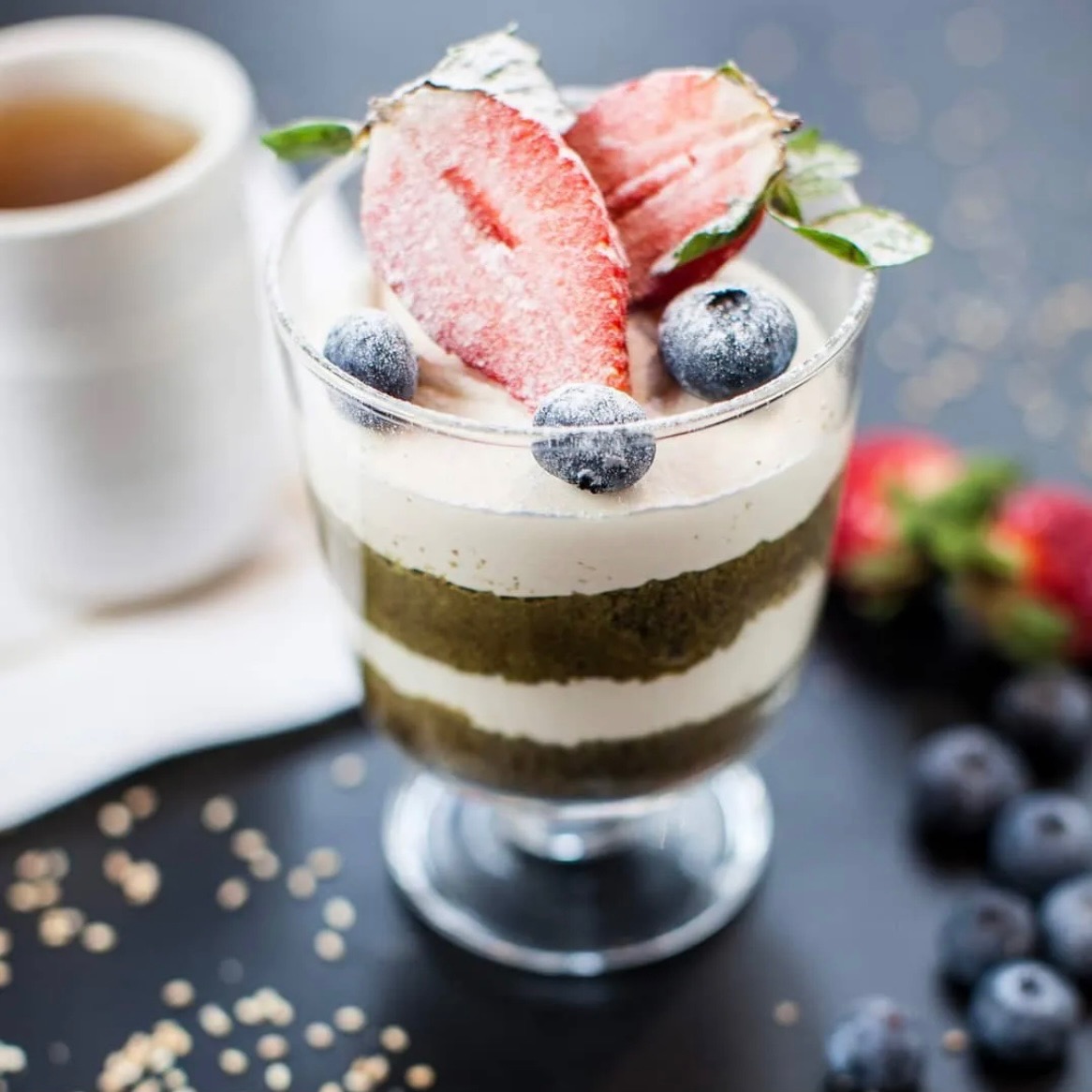 Deser warstwowy matcha z kremem, truskawkami w pudrze i borówkami w szklanym pucharku na ciemnym tle z rozrzuconymi borówkami i kaszą.