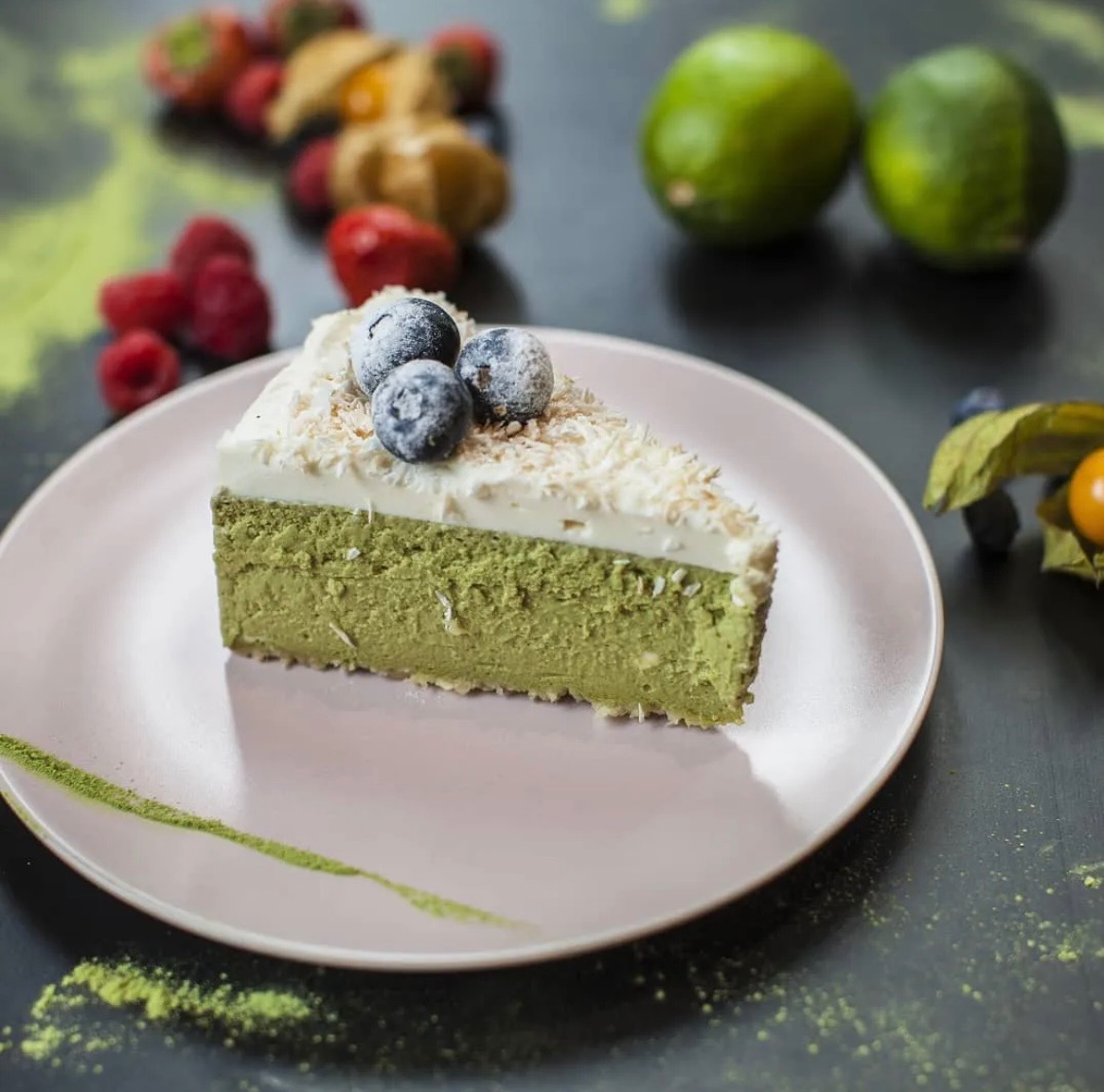 Kawałek zielonego ciasta matcha z kremem, wiórkami kokosowymi i borówkami na różowym talerzu, z owocami i limonkami w tle.