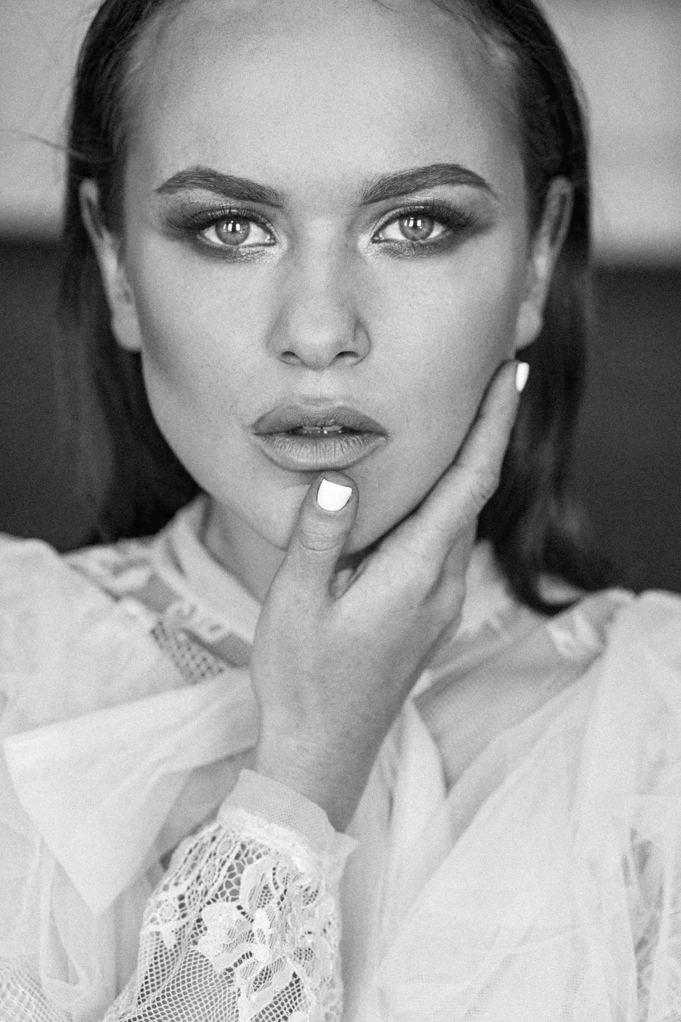 Portret kobiety w czerni i bieli, z dłonią na policzku, subtelnym makijażem oczu i białym manicure, ubrana w delikatną bluzkę z koronkowymi detalami.