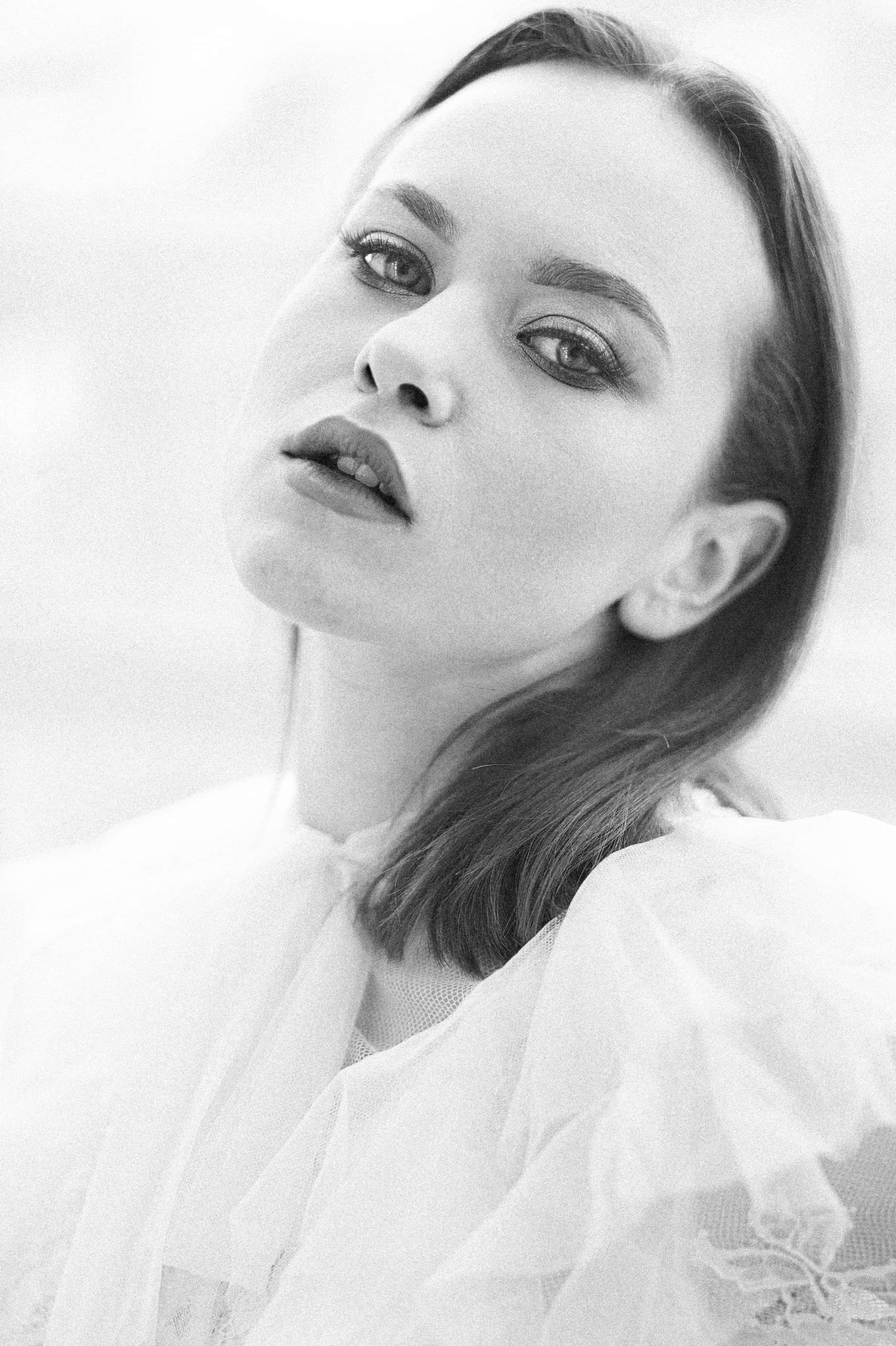 Portret kobiety w białej, zwiewnej sukience, z głową lekko odchyloną do tyłu, w czarno-białej tonacji, z wyraźnym makijażem oczu i ust.