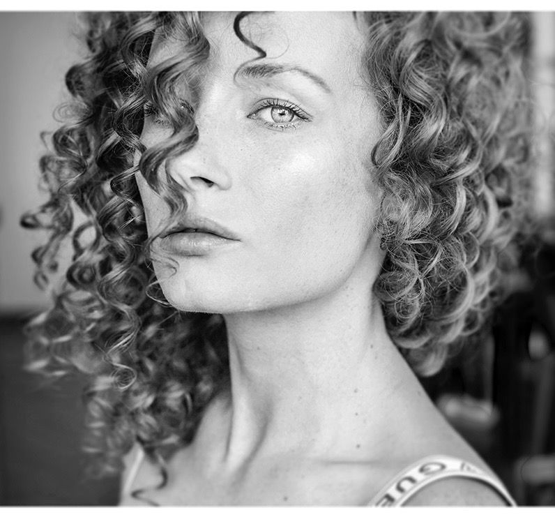 Portret kobiety z burzą kręconych włosów częściowo zasłaniających twarz, czarno-białe ujęcie z bliska, skupione na wyrazistym spojrzeniu.