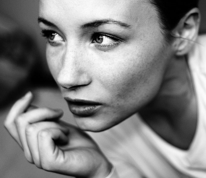 Portret kobiety w czerni i bieli, zamyślony wyraz twarzy, delikatne światło podkreśla rysy, dłoń w pobliżu ust, miękkie tło.