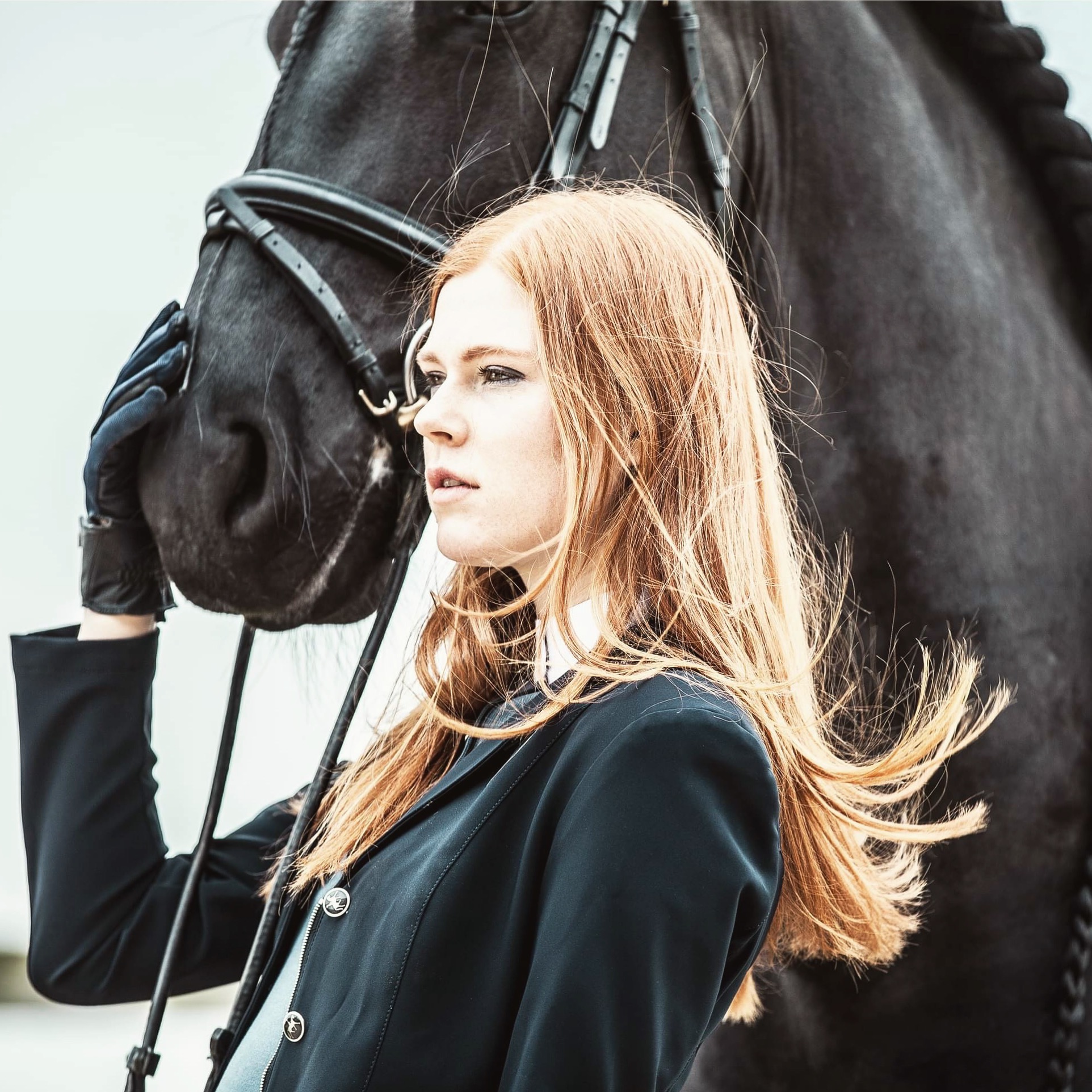 Portret kobiety o rudych włosach w czarnym żakiecie, trzymającej uzdę czarnego konia w Warszawie. Kobieta patrzy w dal, a na jej twarzy maluje się spokój.