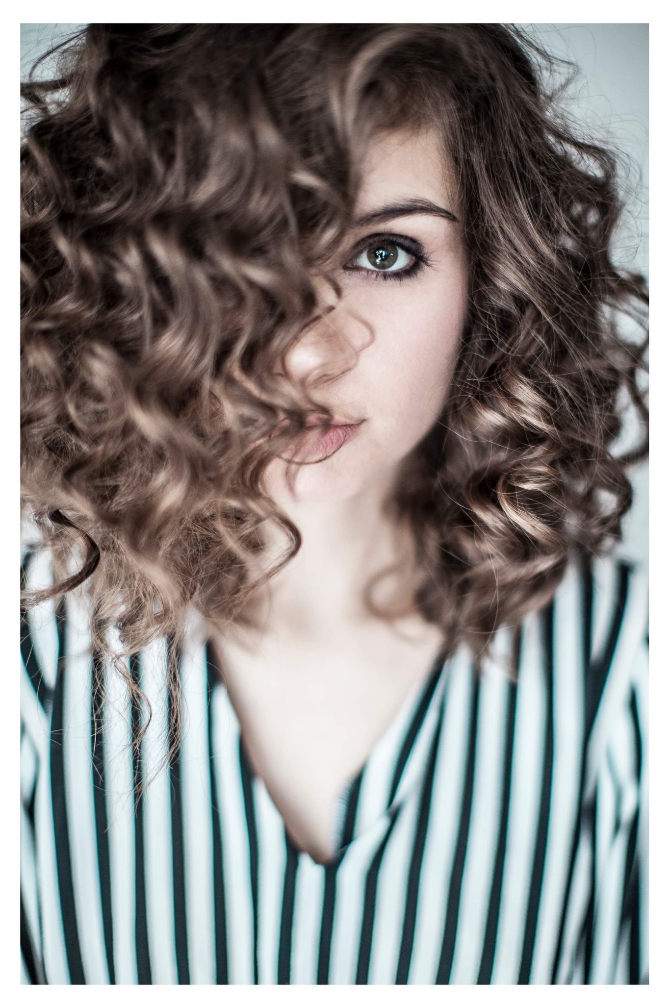 Portret kobiety z kręconymi włosami zasłaniającymi część twarzy, jedno oko widoczne, ubrana w bluzkę w czarno-białe paski.