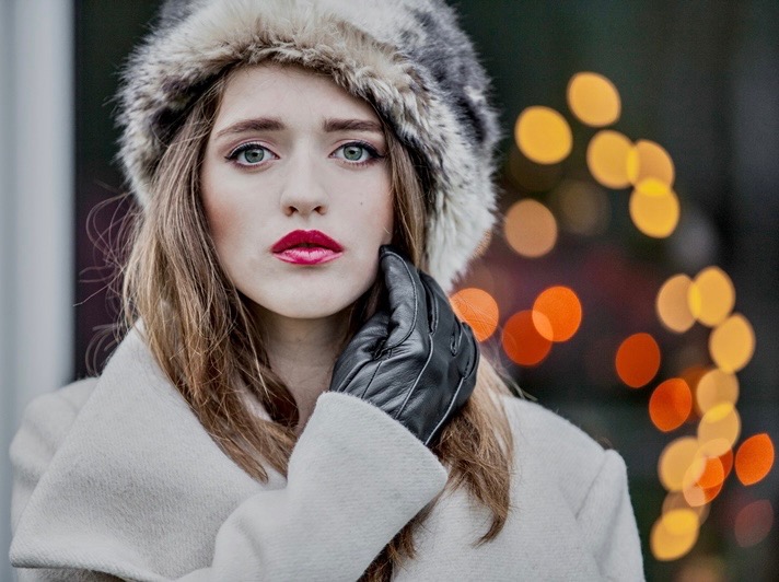 Portret kobiety w futrzanej czapce, białym płaszczu i skórzanych rękawiczkach, z rozmazanym tłem światełek.