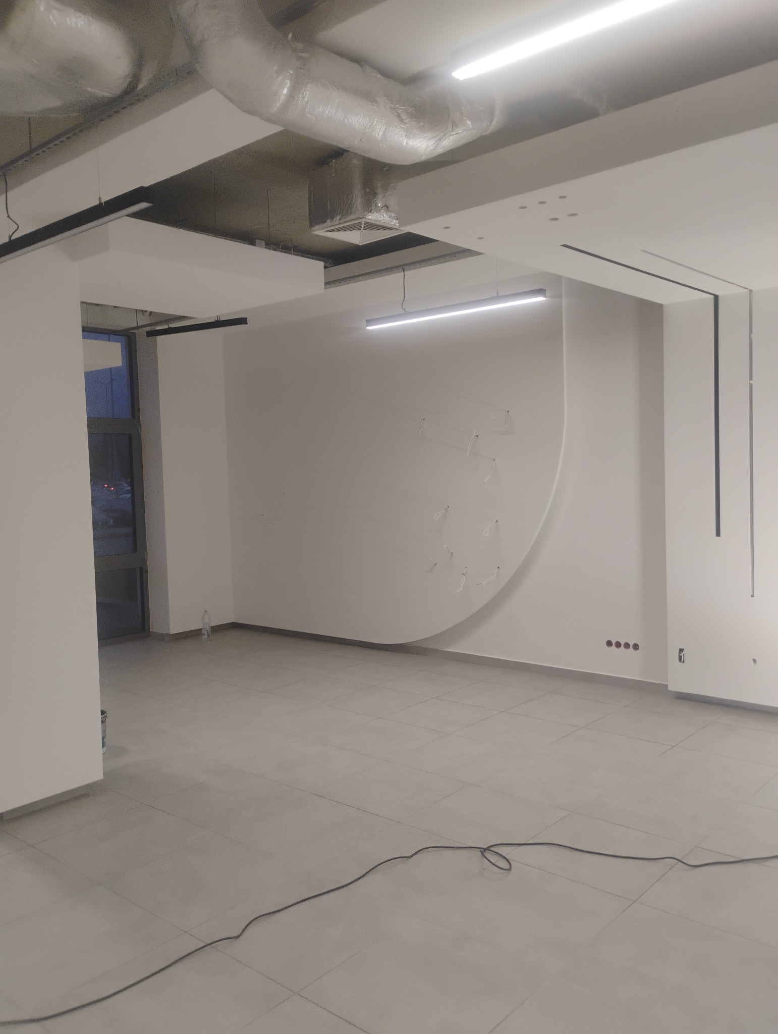 Surowe wnętrze biurowe z szarą podłogą z płytek, białymi ścianami z subtelnymi elementami dekoracyjnymi i widocznymi instalacjami pod sufitem, w tym wentylacja i oświetlenie liniowe.