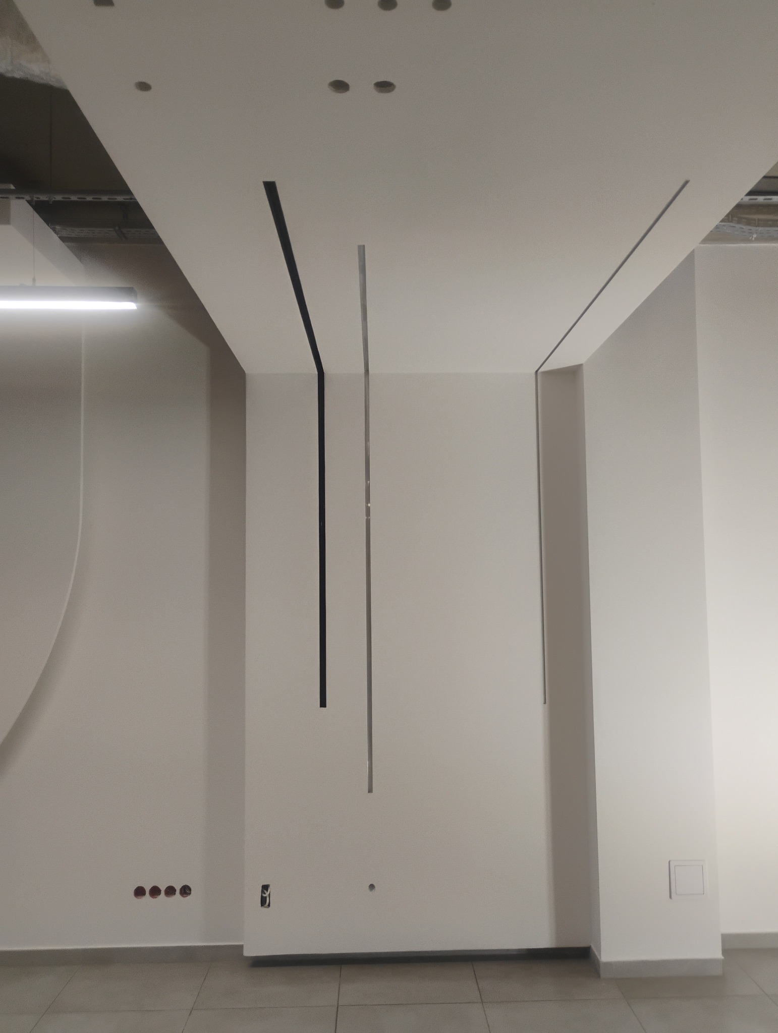 Nowoczesne, minimalistyczne wnętrze biurowe z białymi ścianami i sufitem podwieszanym, ozdobione pionowymi, wpuszczanymi listwami oświetleniowymi w kolorze czarnym i szarym.