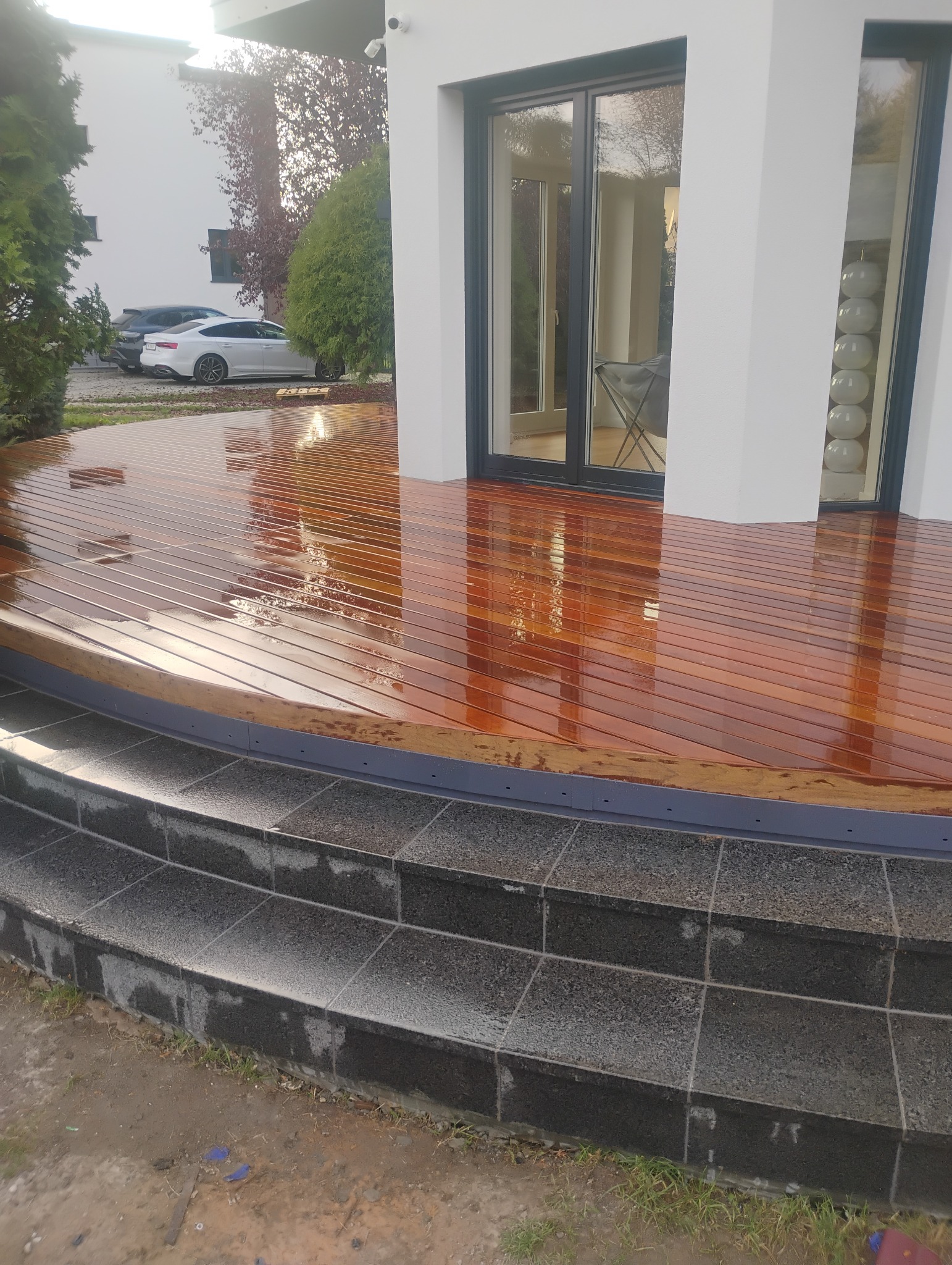 Świeżo polakierowany, zakrzywiony taras z egzotycznego drewna prowadzący do nowoczesnego domu z dużymi oknami przesuwnymi, widoczne kamienne schody.