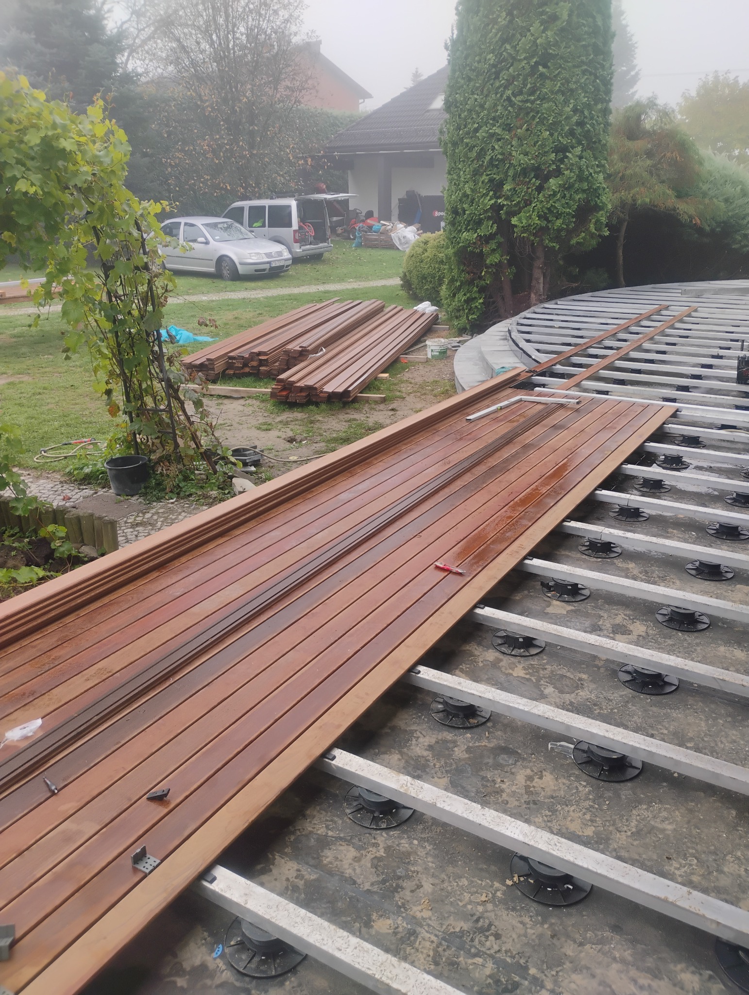 Montaż drewnianego tarasu na regulowanych wspornikach; widoczne deski tarasowe, aluminiowa konstrukcja nośna i stos desek w tle, dom jednorodzinny i samochody w oddali.