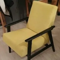 Żółty fotel z czarnymi drewnianymi podłokietnikami i nogami, w stylu retro, wykonany w Olsztynie. Widoczne niedoskonałości oświetlenia i tła sugerują ręczną pracę.