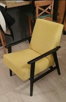 Żółty fotel z czarnymi drewnianymi podłokietnikami i nogami, w stylu retro, wykonany w Olsztynie. Widoczne niedoskonałości oświetlenia i tła sugerują ręczną pracę.