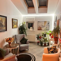 Psycholog Edyta Tabor - Centrum Zintegrowanego Wsparcia Psychicznego MindsET - Przytulne wnętrze gabinetu z dwoma fotelami w pepitkę i jednym kremowym, okrągły czarny dywan z białymi paskami, liczne rośliny doniczkowe, obrazy na ścianach i okna dachowe.