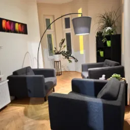 Przytulne wnętrze gabinetu z trzema szarymi fotelami, lampą łukową i roślinami doniczkowymi na tle jasnych ścian i drewnianej podłogi.