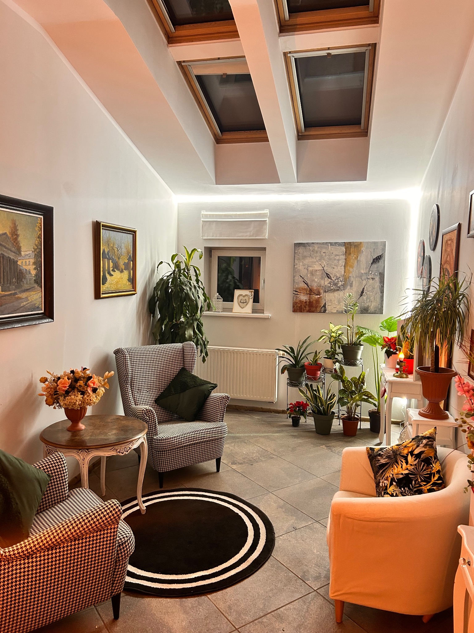 Przytulne wnętrze gabinetu z dwoma fotelami w pepitkę i jednym kremowym, okrągły czarny dywan z białymi paskami, liczne rośliny doniczkowe, obrazy na ścianach i okna dachowe.