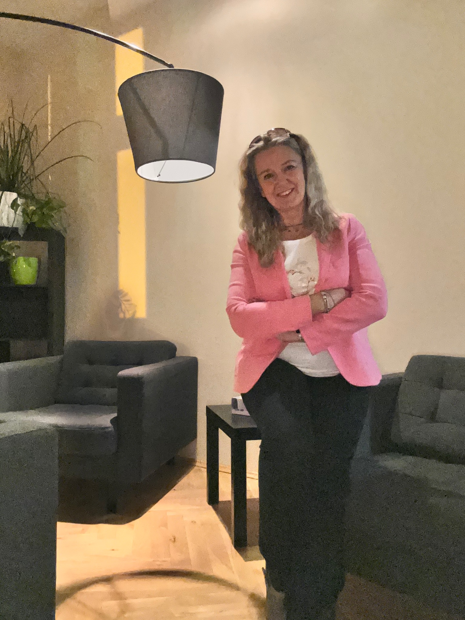 Uśmiechnięta kobieta w różowej marynarce, stojąca w przytulnym gabinecie z szarymi fotelami i lampą łukową. W tle widoczna półka z roślinami.