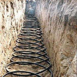 P.W.Eko-Live J.Krzysztofczyk - Wykop z ułożonym poziomym kolektorem gruntowym pompy ciepła w postaci czarnej rury w kształcie spirali, widoczni pracownicy i koparka w tle.