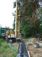 Wiertnica zamontowana na ciężarówce, przygotowana do wiercenia studni głębinowej na zalesionej działce, widoczne węże i elementy instalacji.