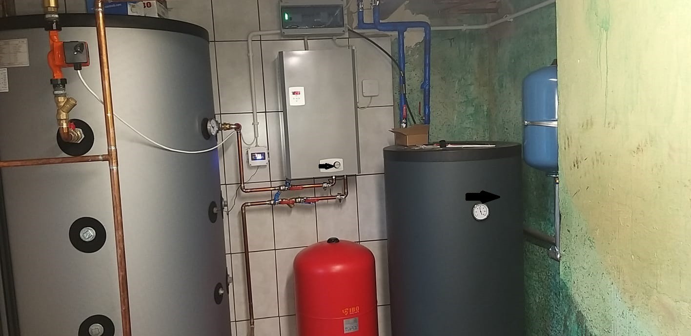 Kotłownia z dwoma szarymi zbiornikami akumulacyjnymi, czerwonym zbiornikiem wyrównawczym, szarym piecem gazowym wiszącym, instalacją z miedzianych rur i niebieskimi rurami z tworzywa, na tle...