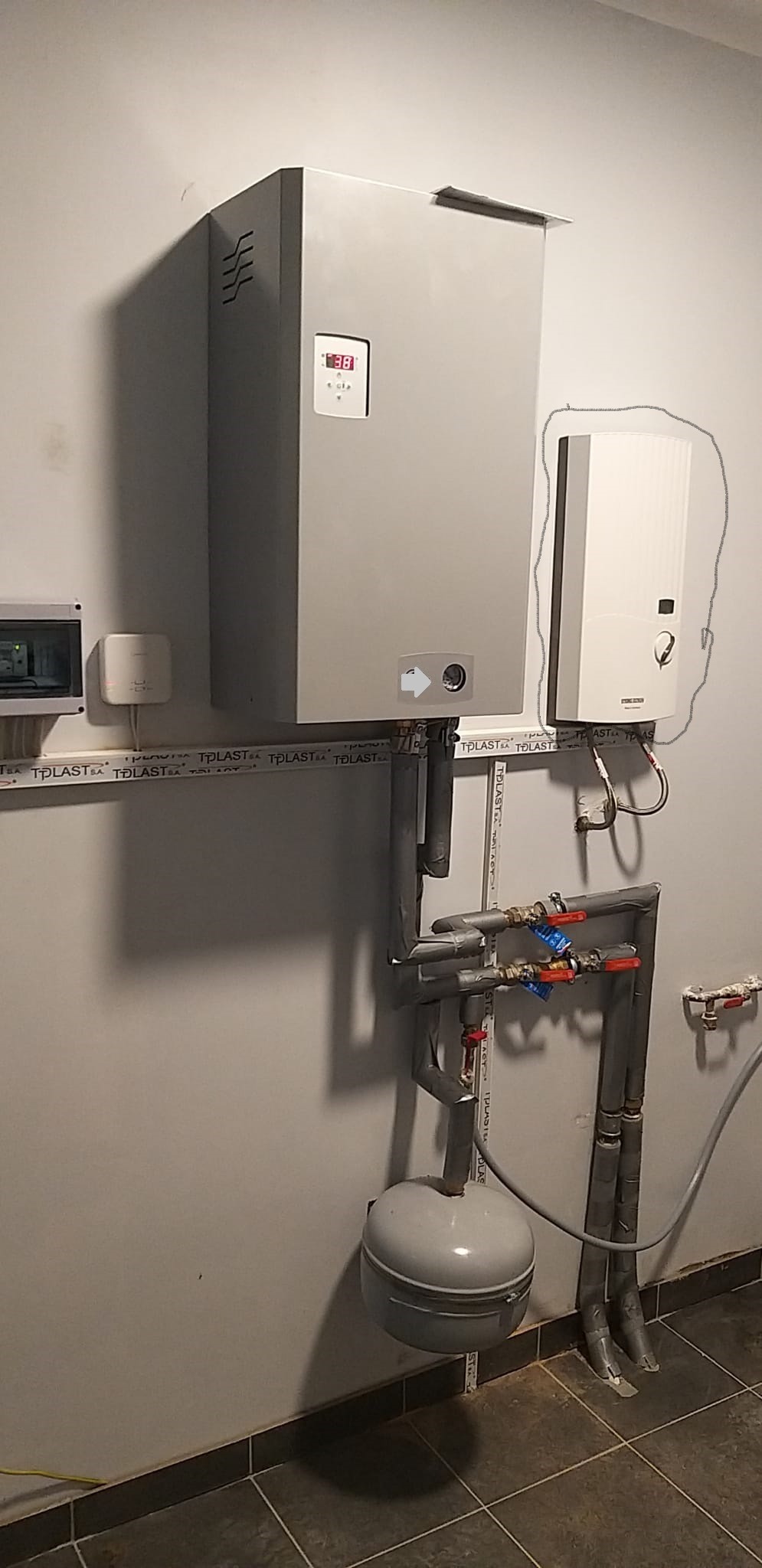 Szara instalacja grzewcza z piecem i zbiornikiem wyrównawczym, rury izolowane szarą taśmą, na ścianie widoczny dodatkowy moduł sterujący.