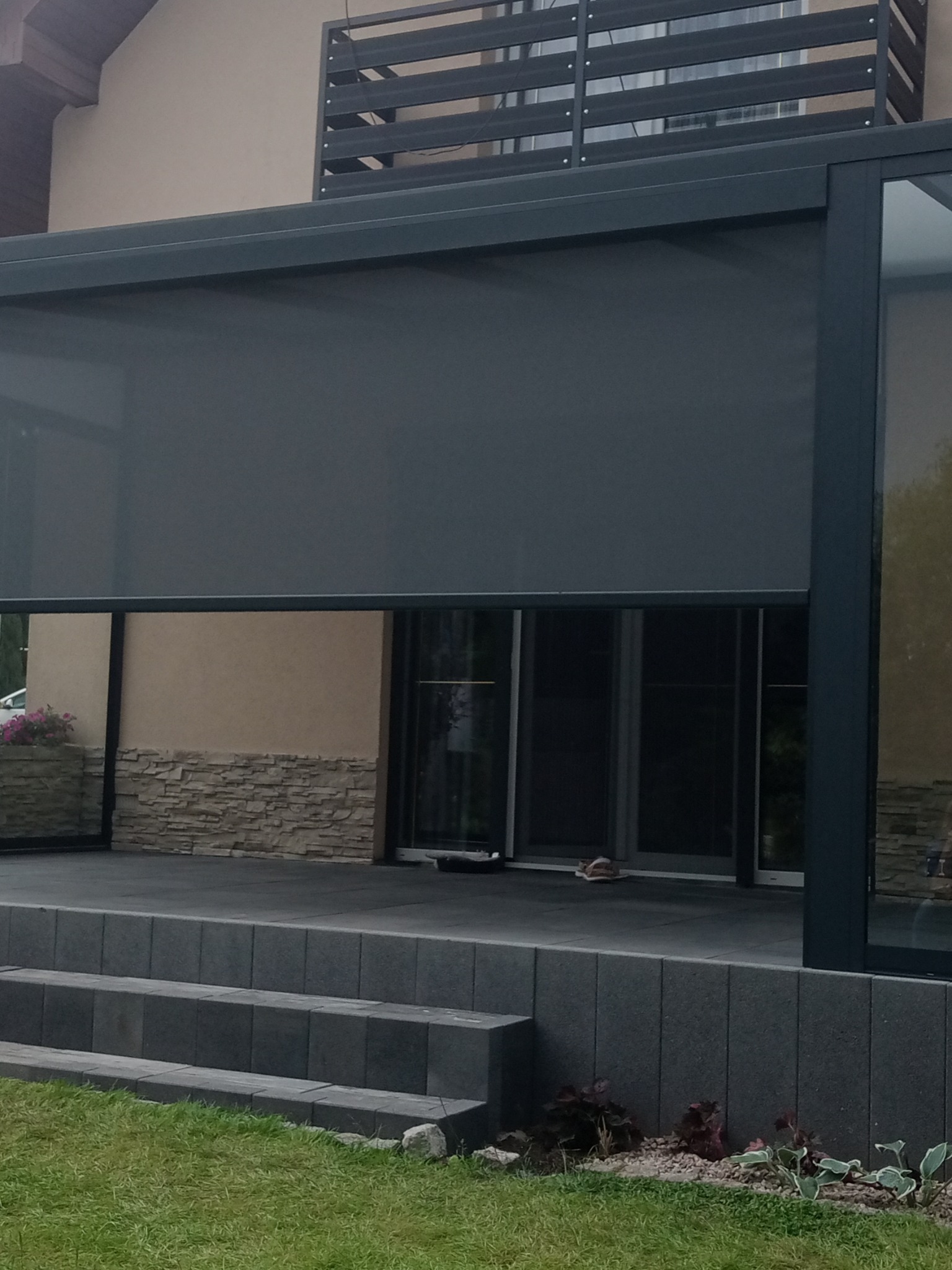 Zewnętrzna, częściowo opuszczona roleta przeciwsłoneczna w kolorze grafitowym, zamontowana przy tarasie z szarych płyt, widoczne schody z kostki brukowej i trawnik.