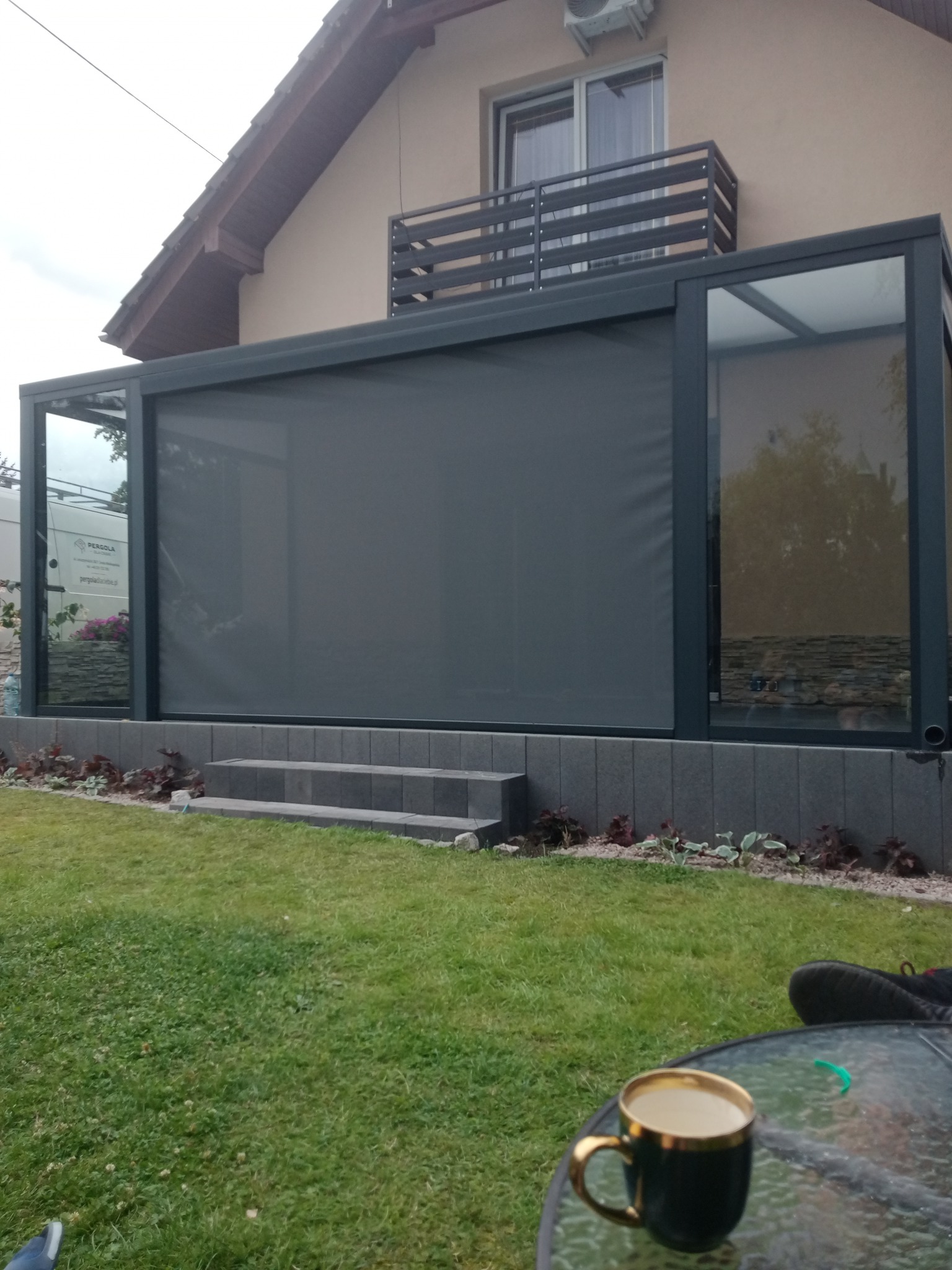 Zadaszony taras z szarą roletą przeciwsłoneczną, widok z ogrodu, filiżanka kawy na szklanym stoliku w pierwszym planie.