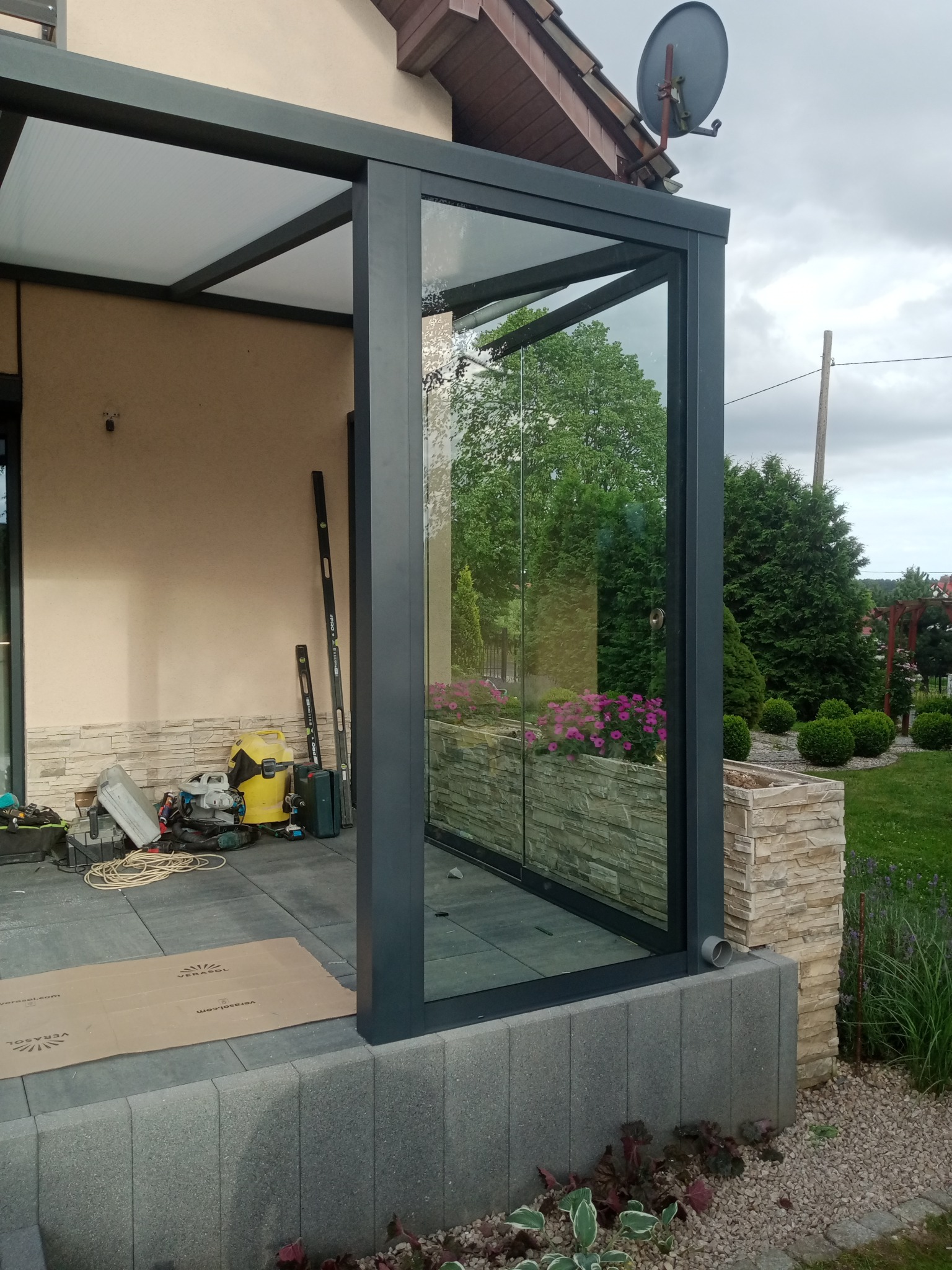 Szklana zabudowa tarasu z przesuwnymi drzwiami, widoczne narzędzia i poziomice, karton ochronny na podłodze, ogród w tle.