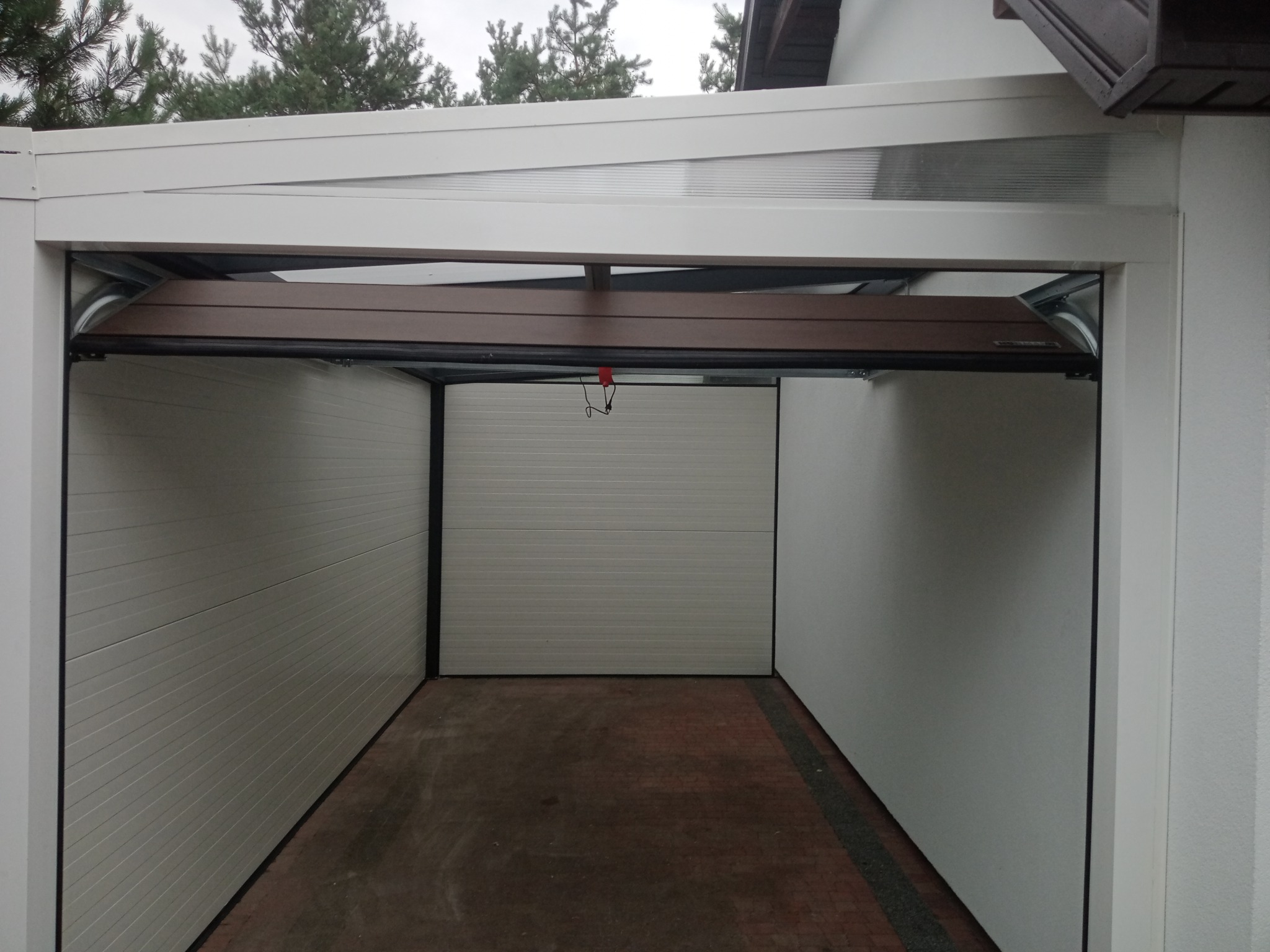 Częściowo otwarta, segmentowa brama garażowa w kolorze brązowym, z widocznym mechanizmem podnoszenia, w garażu z jasnymi ścianami i czerwoną kostką brukową na podłodze.